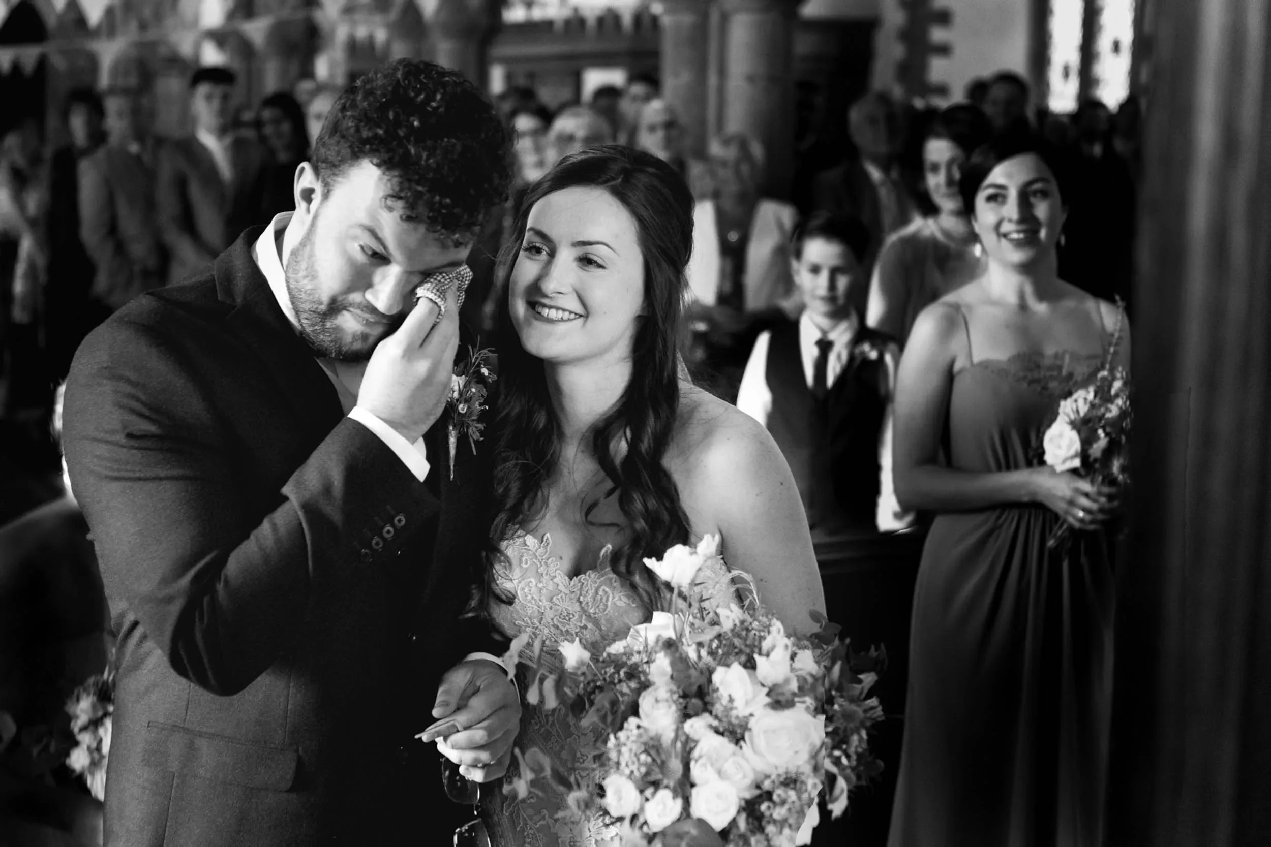 A black and white photograph of a wedding ceremony inside a church. The bride, holding a large bouquet of flowers, is smiling at the groom, who is wiping away tears with a tissue. Bridesmaids and guests stand in the background, observing the moment.