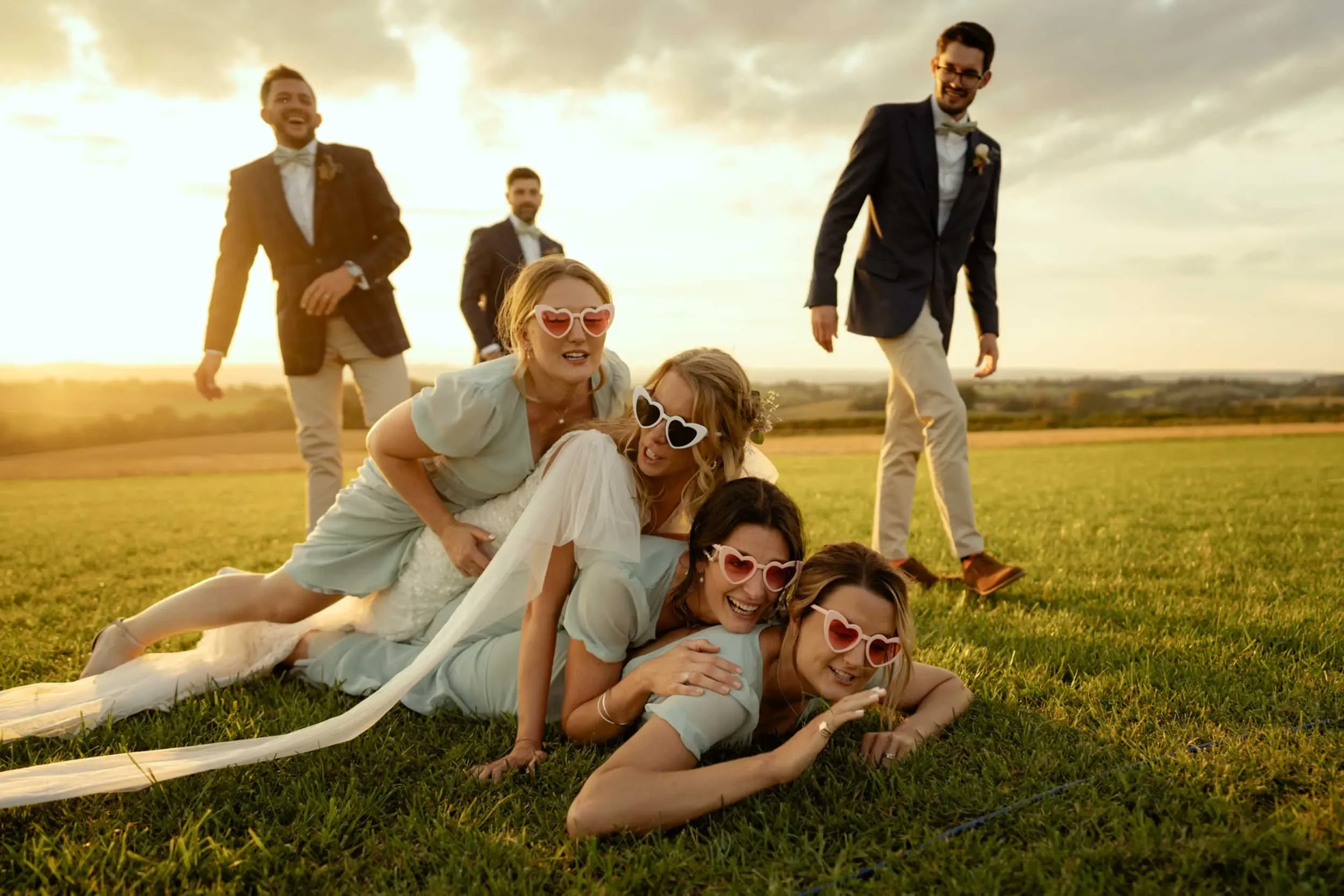 A group of six friends, four women and two men, enjoying a photo on a grassy field during sunset. The women are lying on the ground in a line, smiling and wearing heart-shaped sunglasses, with one woman on top of the others. The men stand in the back