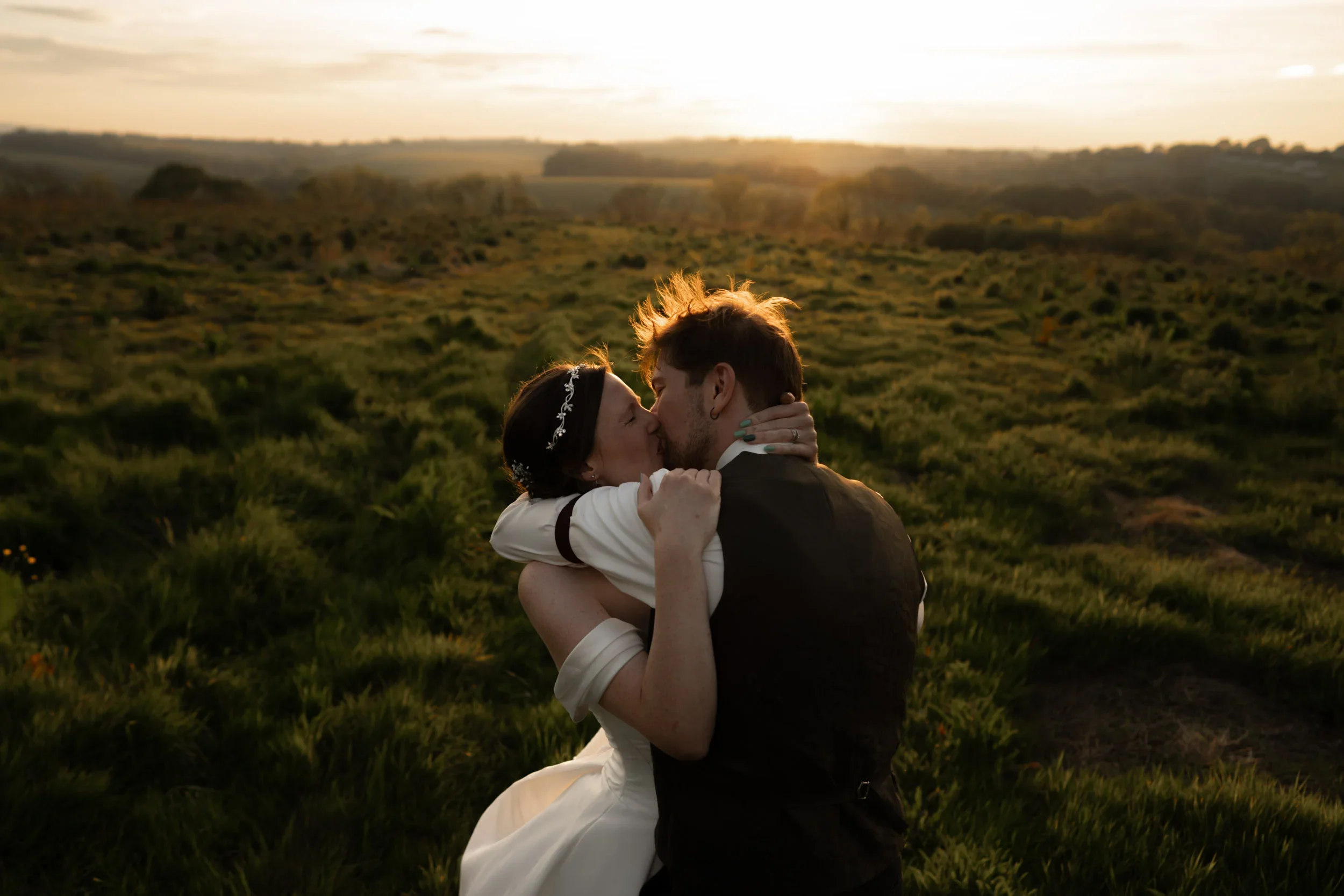 A couple kissing outdoors during sunset, in a green field with rolling hills in the background.