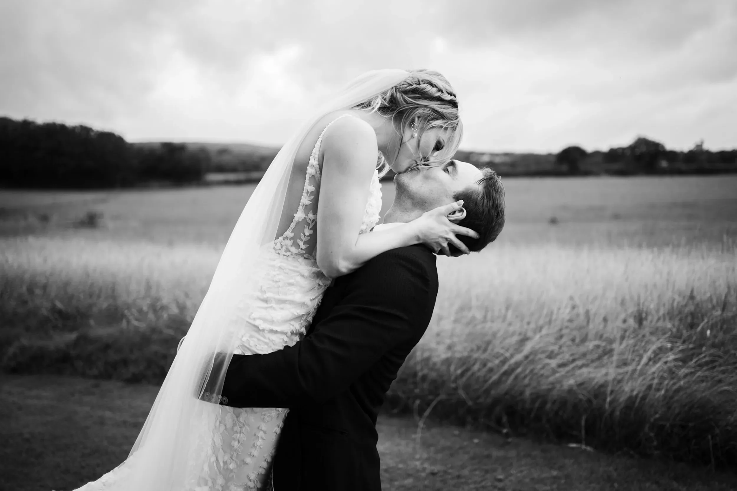 A bride and groom sharing a kiss outdoors in a field, with the bride being lifted by the groom, in black and white.