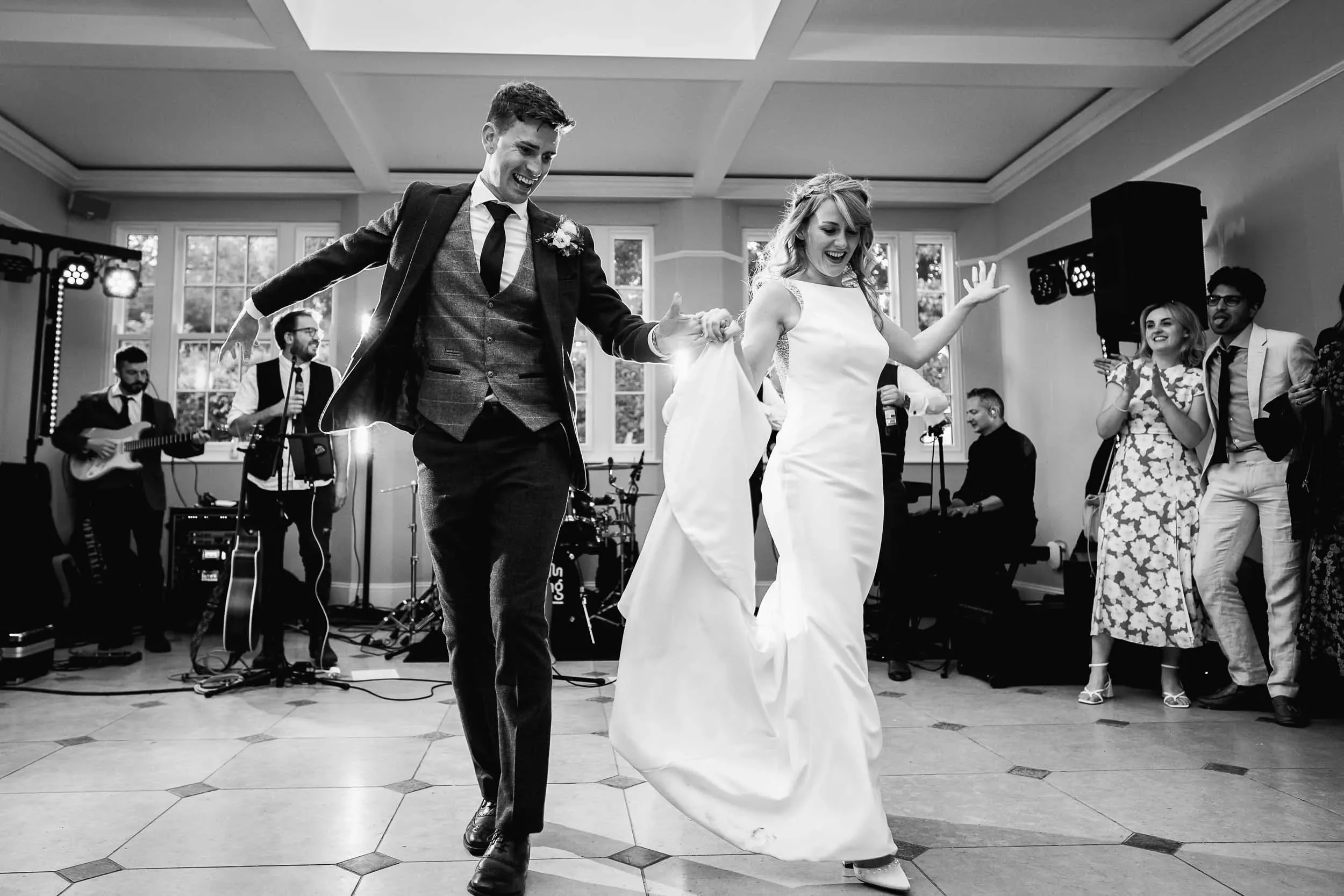 Black and white photo of a bride and groom dancing at their wedding reception, with a band playing in the background and guests watching.