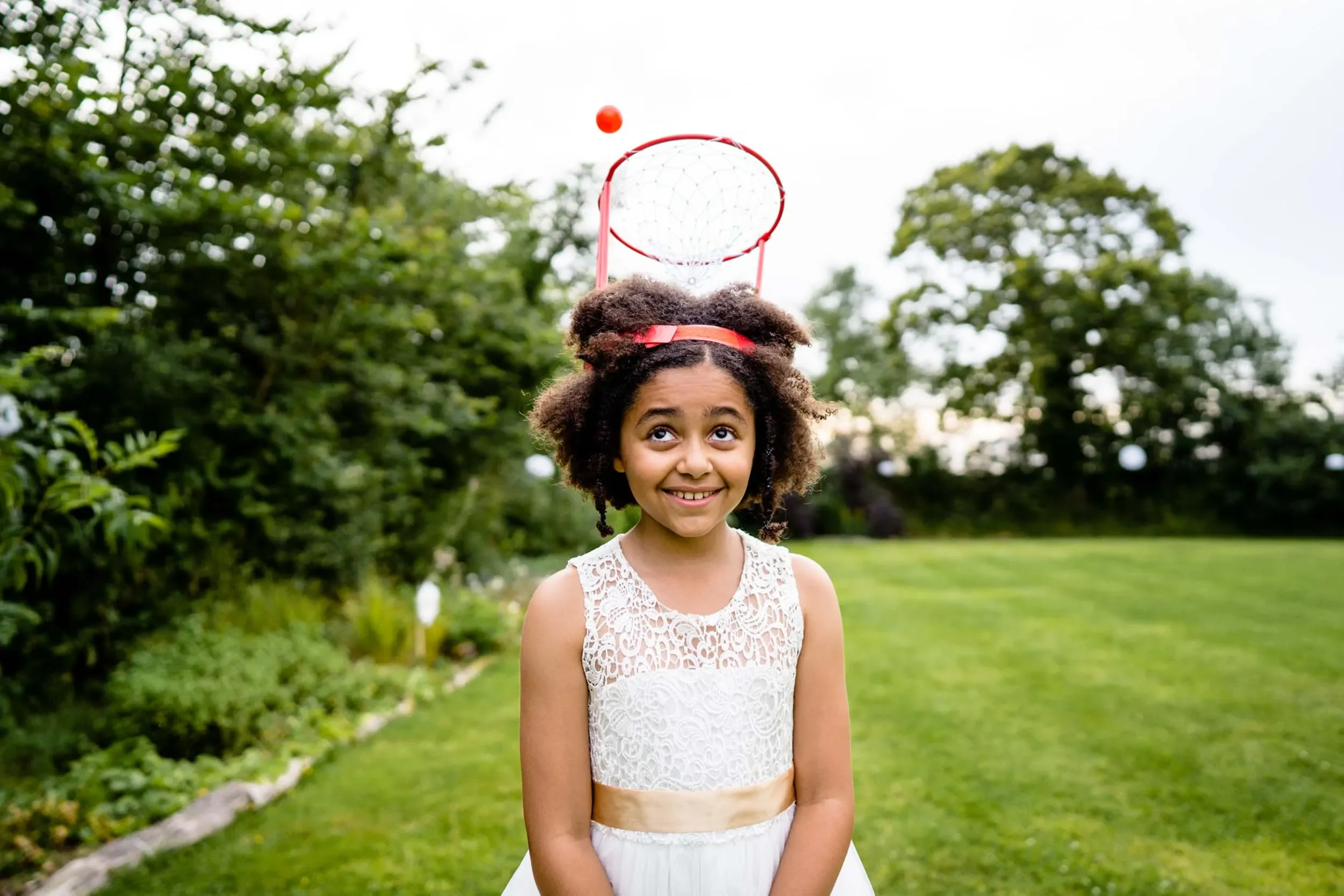 A young girl with curly hair smiling outdoors, wearing a white lace dress with a beige sash, balancing a small basketball hoop on her head, with a basketball in the air.