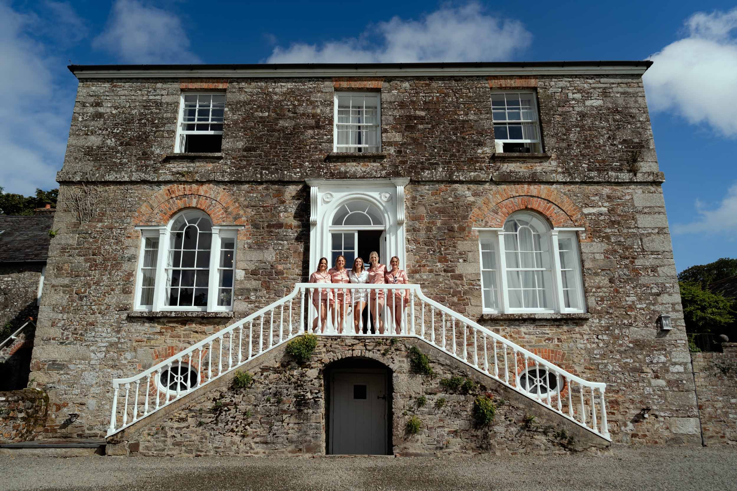 A large, old brick house with four windows on the upper floor and four arched windows on the lower floor. Four women in matching pink satin pajamas stand on the front porch, which is accessed by a white staircase.