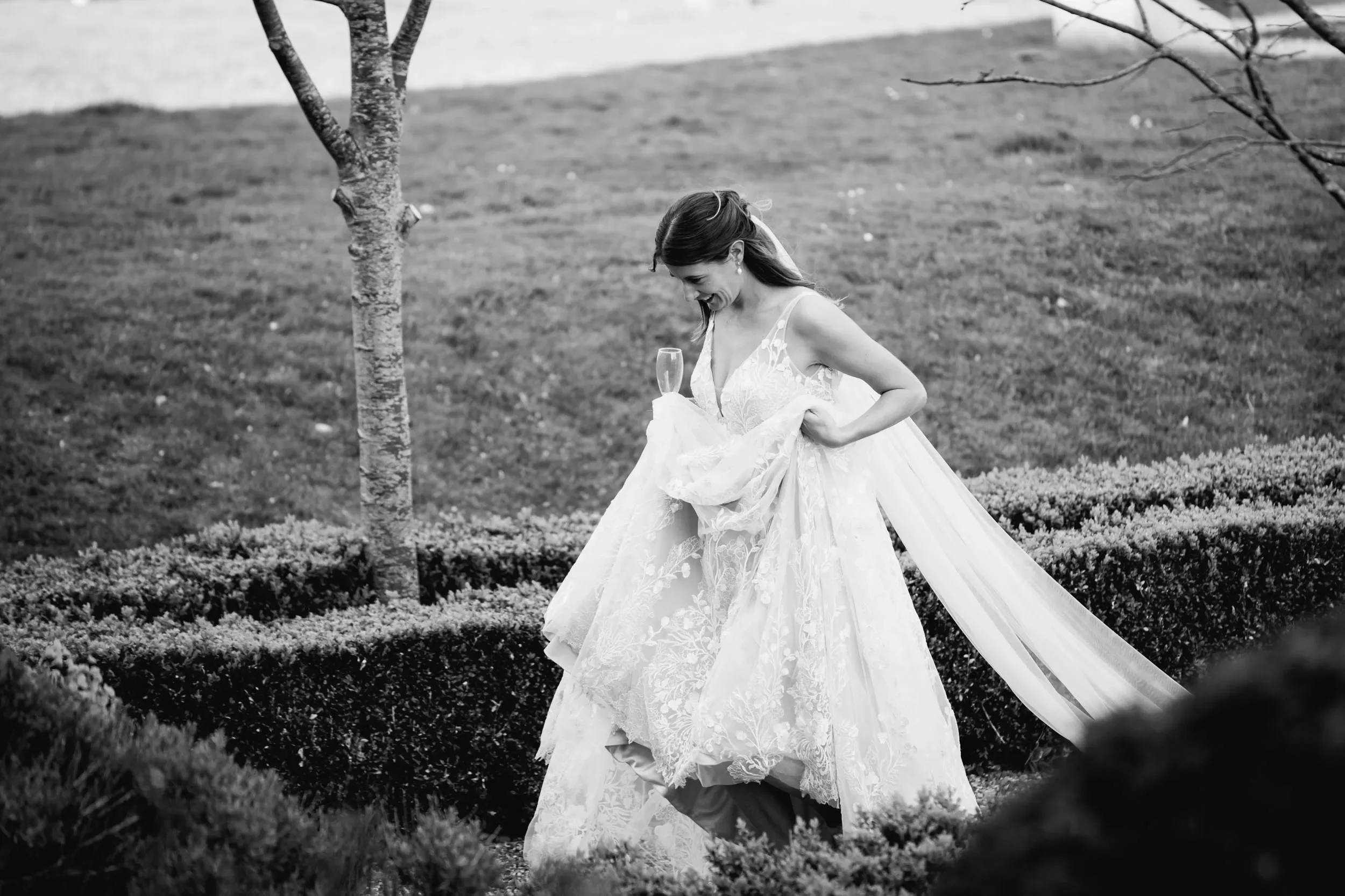 A bride in a wedding dress holding a champagne glass in huntsham court garden with trees and bushes