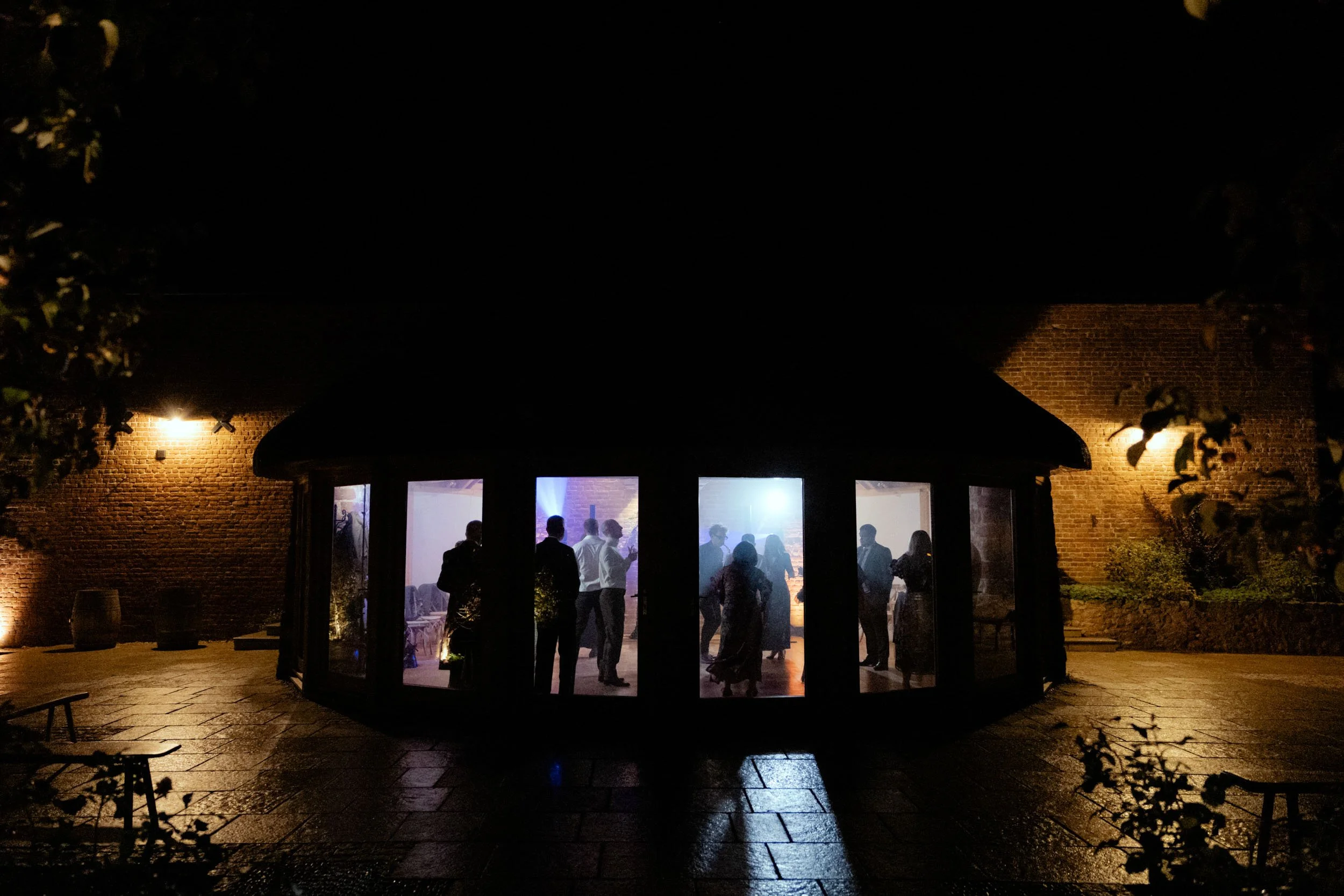 People inside a glass-enclosed room at night, with some socializing and dancing, illuminated by colorful lights, surrounded by a brick building and outdoor area.