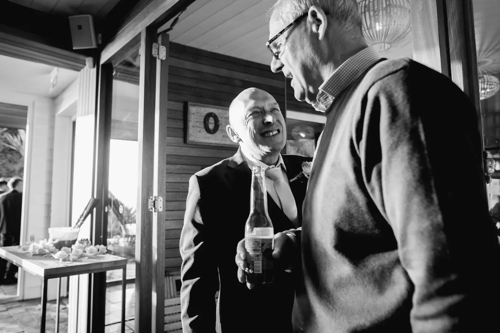 Two men, one in a suit and tie holding a soda bottle, are engaged in a heated conversation at a social gathering. The scene appears to be indoors with wooden walls.