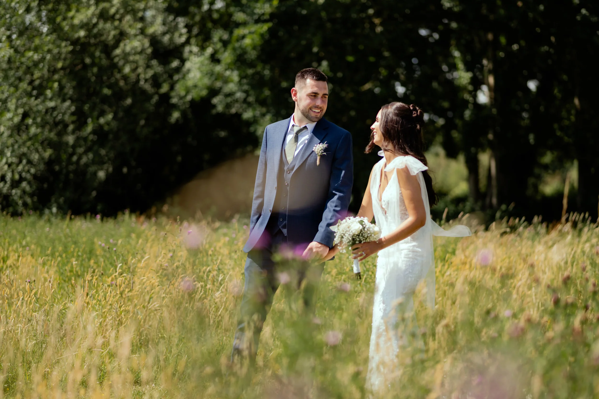 A newlywed couple holding hands and smiling at each other in a grassy field with trees in the background