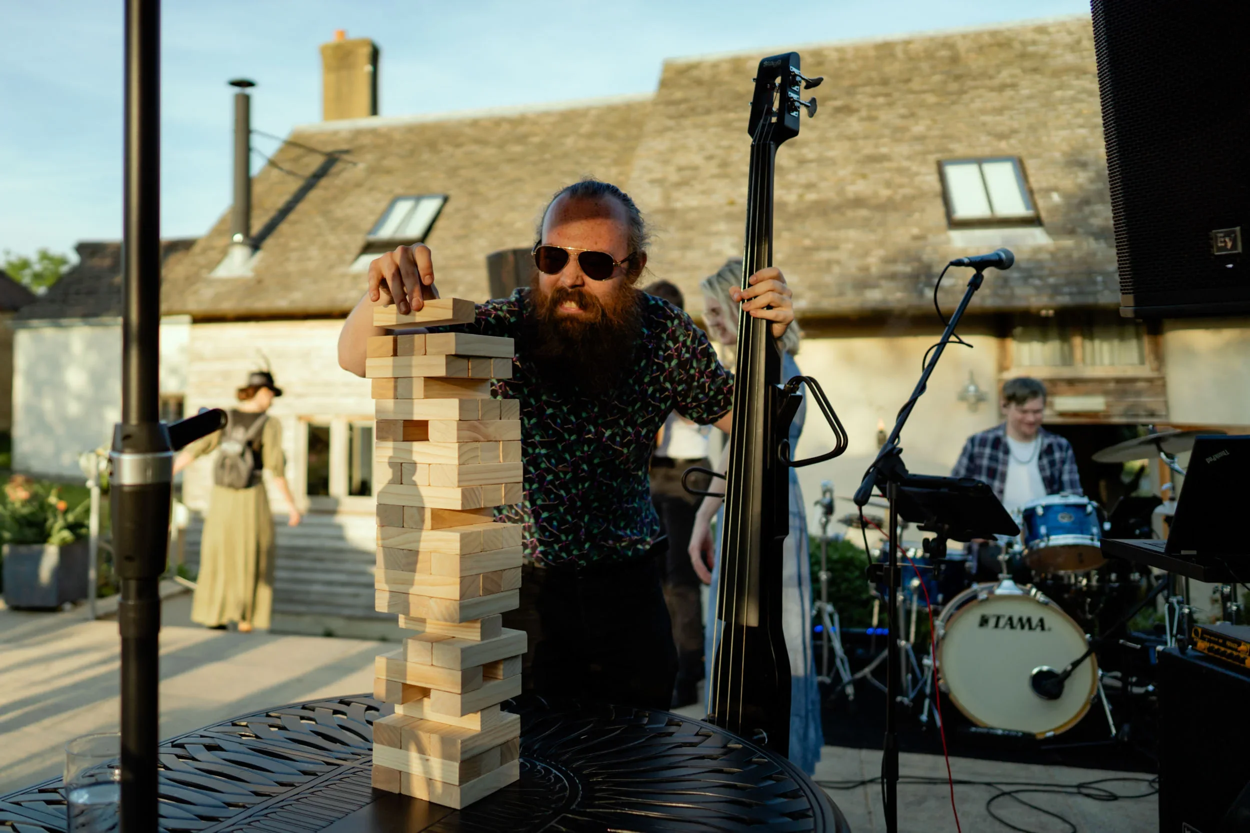 A man with sunglasses and a beard playing outdoor Jenga with large wooden blocks while musicians perform with drums and a double bass. A woman in a long dress and a person in a plaid shirt are in the background, and a house with a stone roof is visib