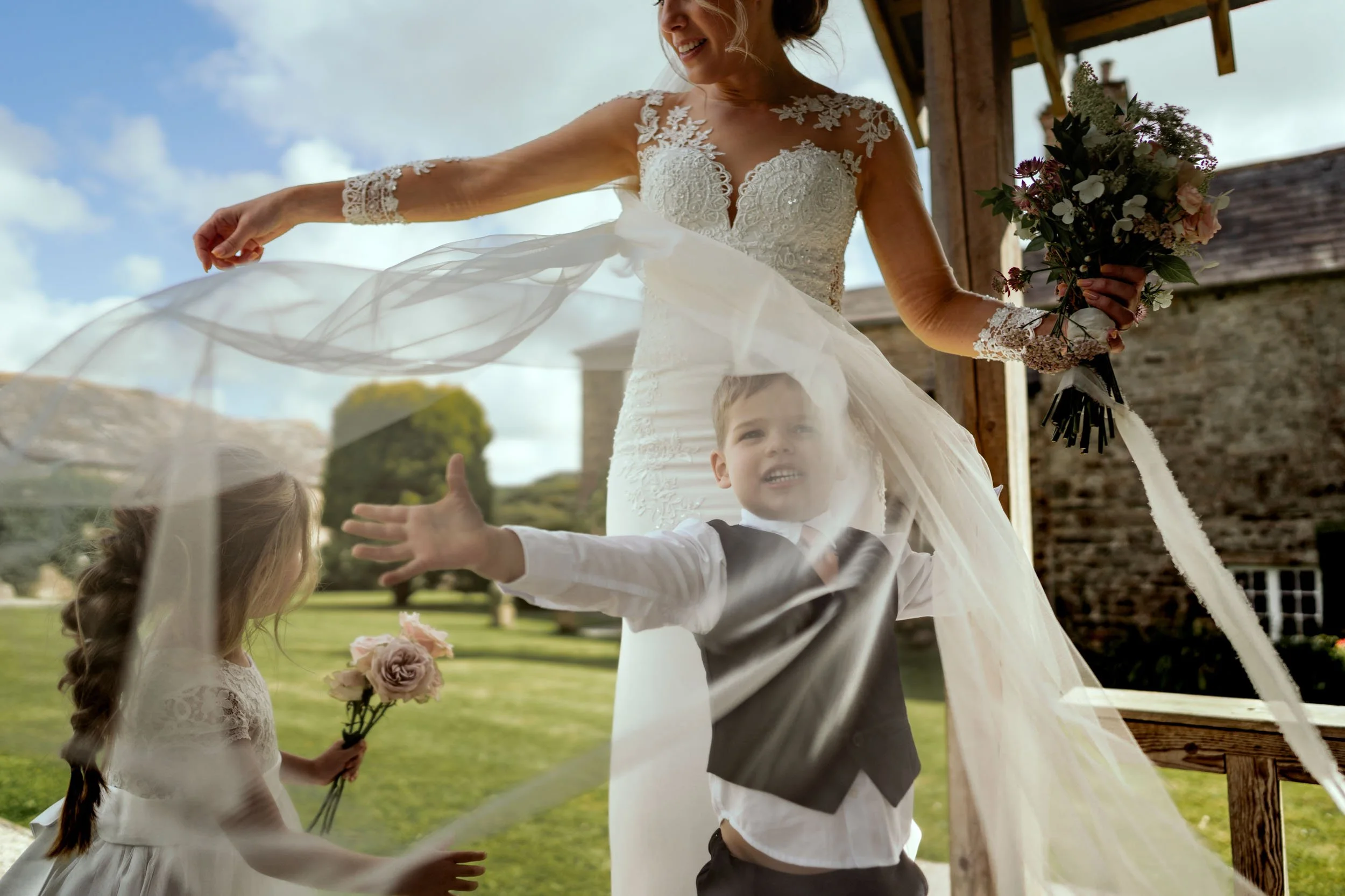 A bride in a wedding dress holding a bouquet of flowers, with two children outside in a garden, one girl holding a small bouquet and one boy reaching out.