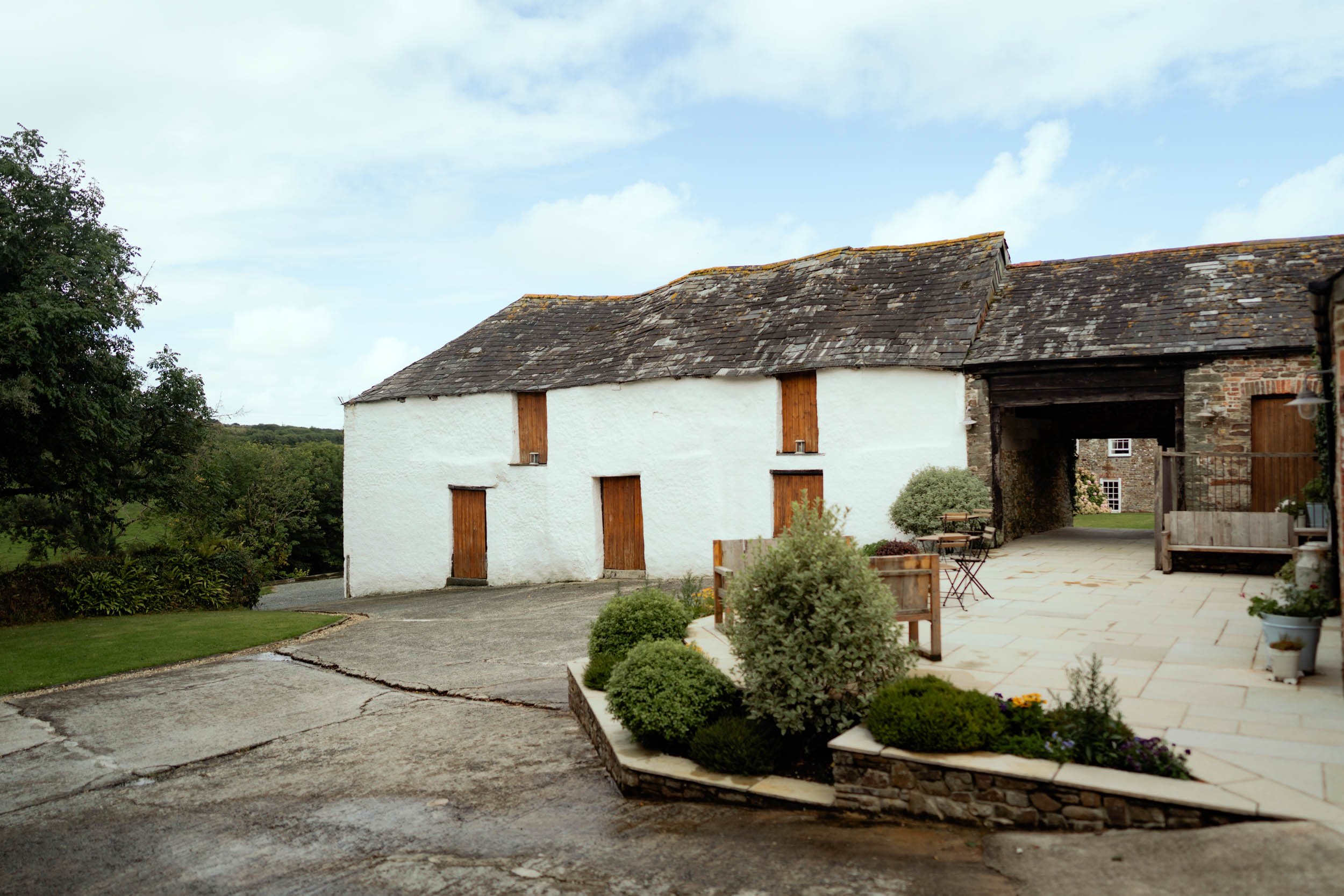 A rustic countryside scene with a white building, wooden shutters, a stone-paved area, and greenery under a partly cloudy sky.
