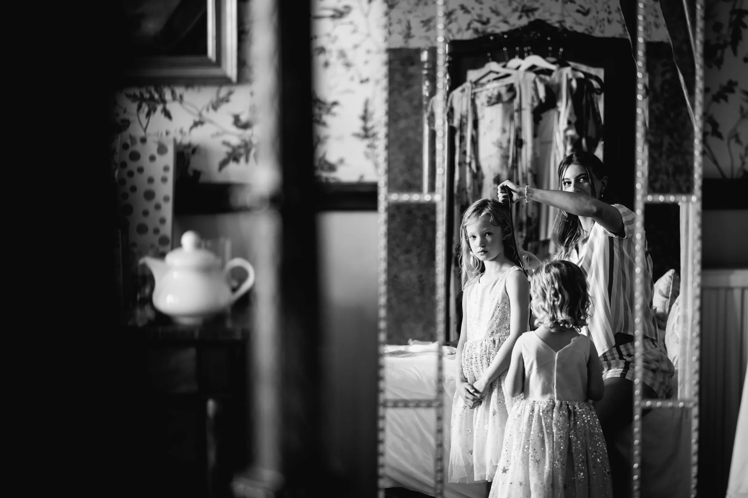 A woman helps two young girls get ready, looking in a mirror in a bedroom with floral wallpaper and hanging clothes.