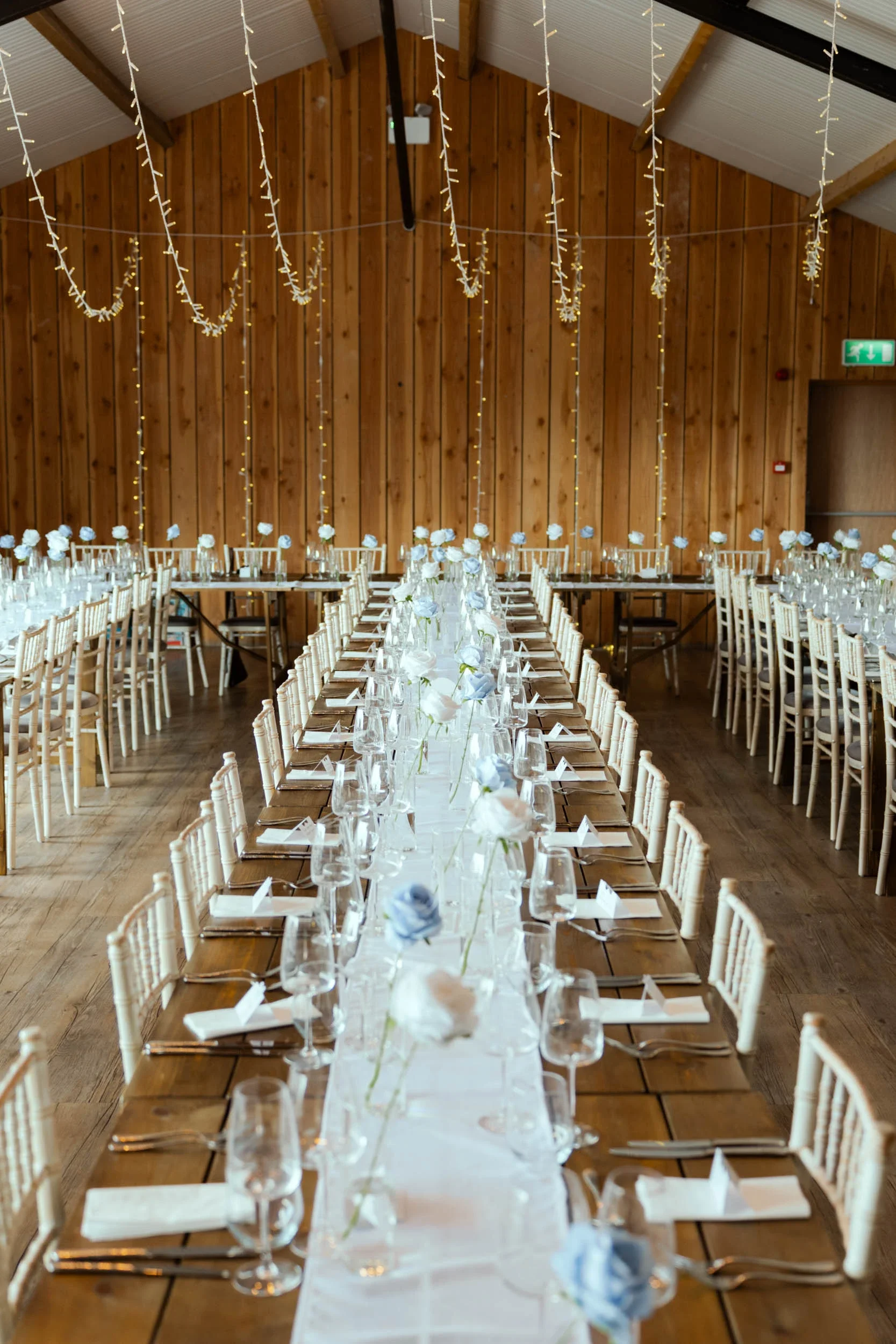 Wedding reception with long central table decorated with white and blue flowers, set with wine glasses, plates, and silverware, surrounded by wooden chairs, inside a wooden venue with hanging string lights.
