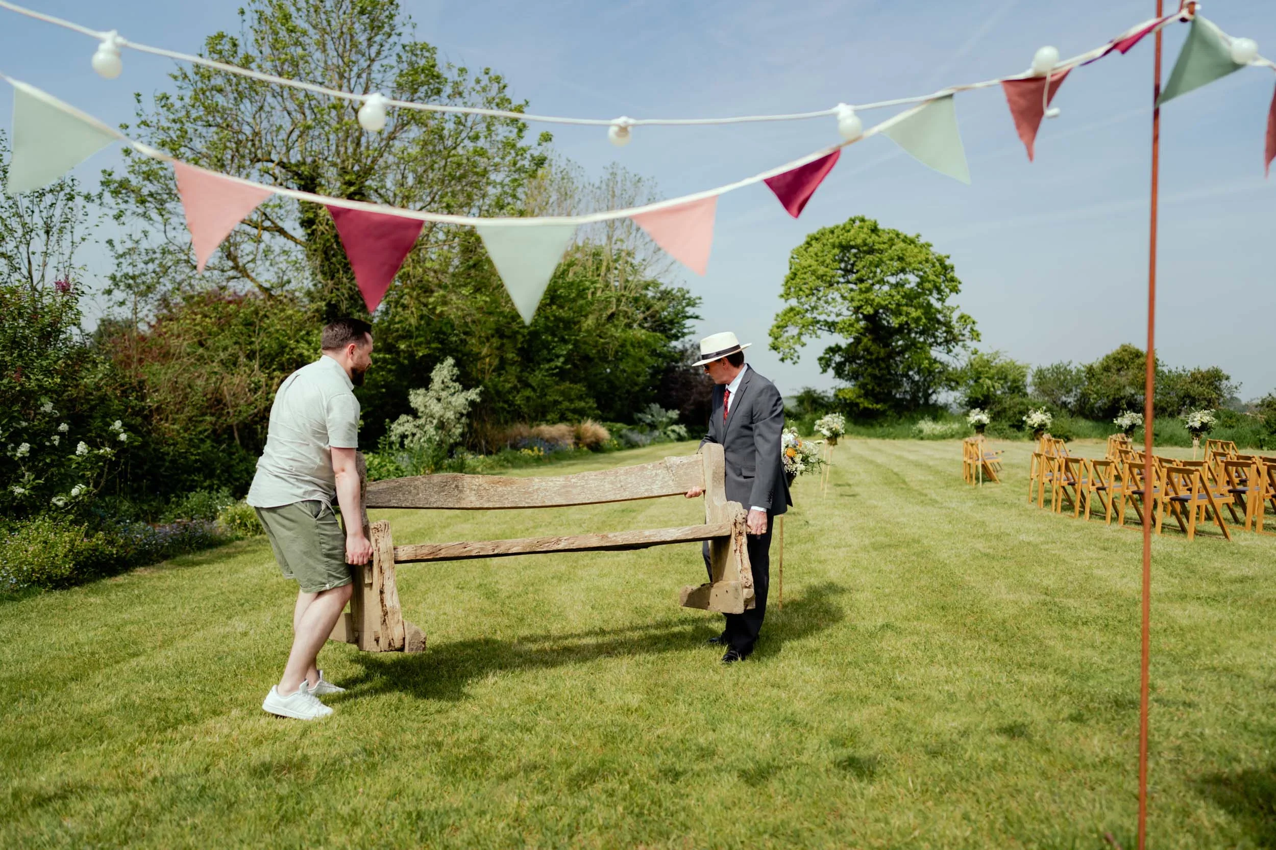 Two men working together to set up a rustic wooden bench outdoors on a sunny day for an event, with chairs and floral arrangements in the background, decorated with colorful bunting and string lights, under a clear sky.