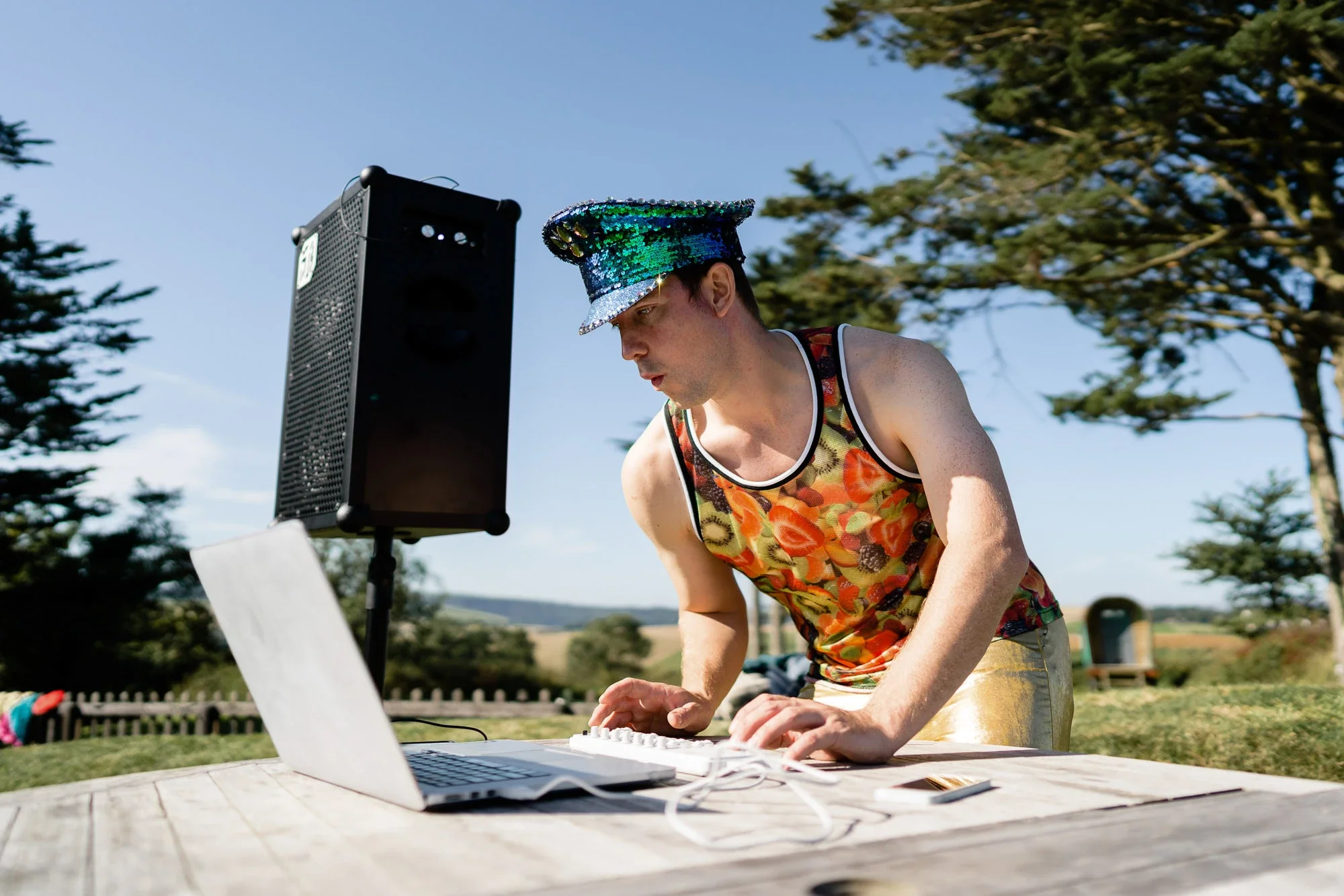 A man in a colorful floral tank top and a shiny, sequin hat is outdoors, working on a laptop connected to a speaker on a wooden table. The background features trees and a clear blue sky.