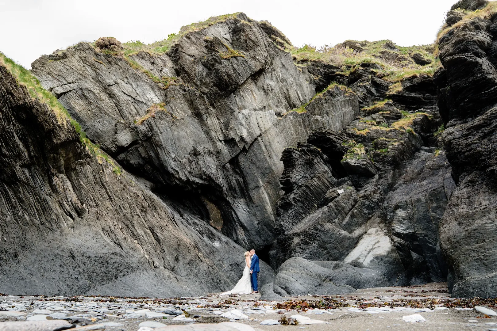A bride and groom stand close together on rocks at a beach, with large, rugged cliffs towering behind them.