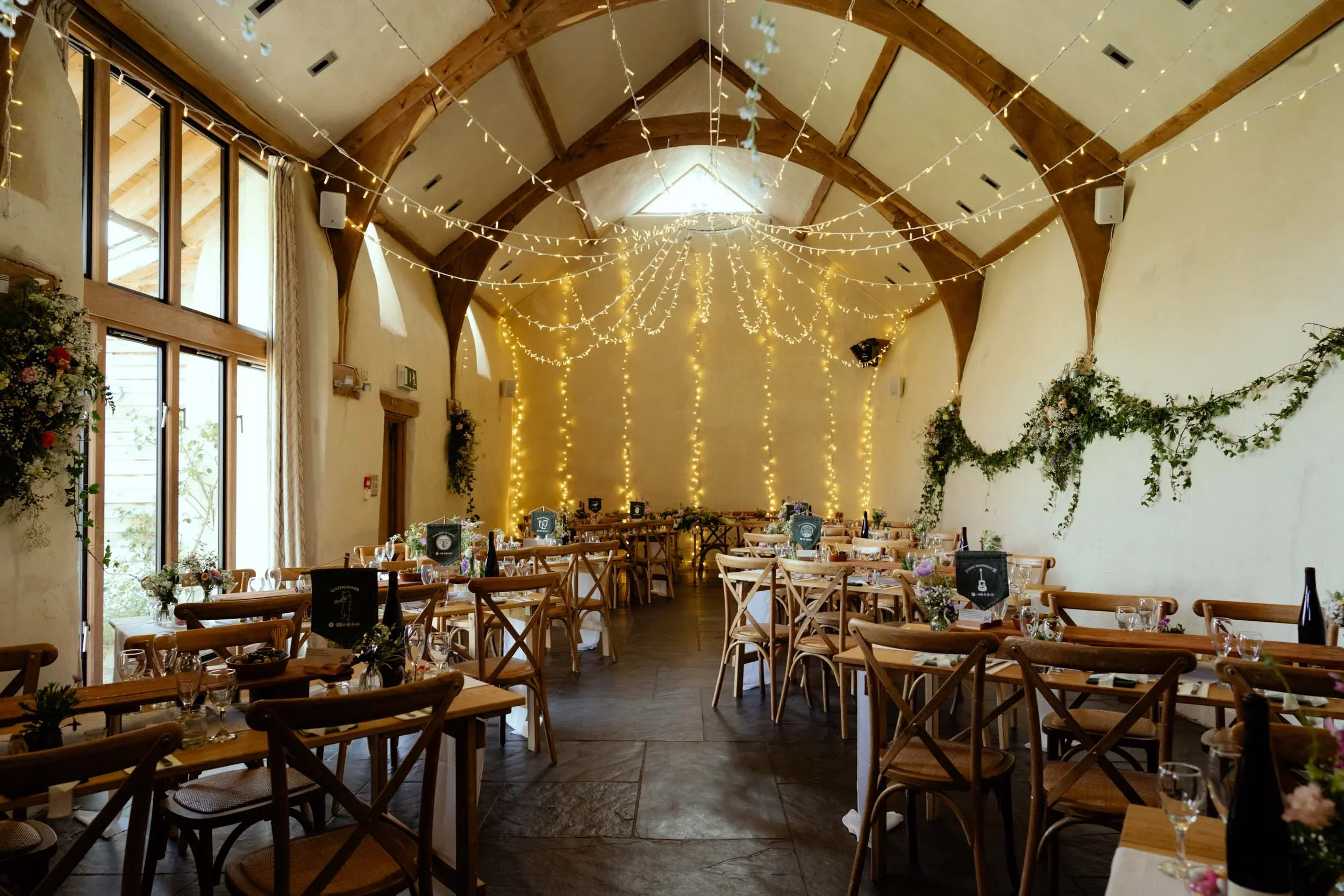 A decorated banquet hall with wooden tables and chairs, string lights hanging from the ceiling, floral arrangements on the walls and tables, and bottles of wine on the tables, set for an event or celebration.
