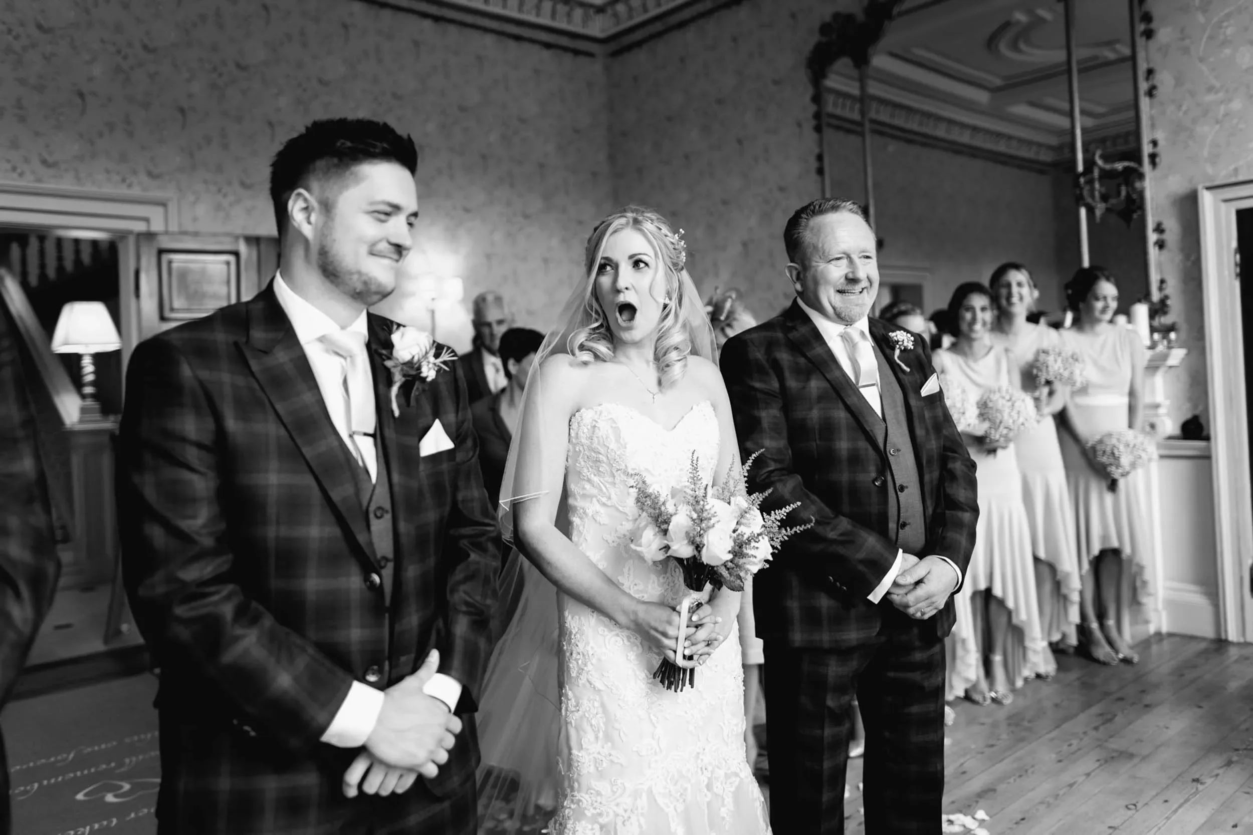 A black-and-white photo of a wedding scene. The bride is in the center, holding a bouquet, with her mouth open and eyes wide. To her right is a man smiling, and to her left is another man with a slight smile. In the background, there are bridesmaids 