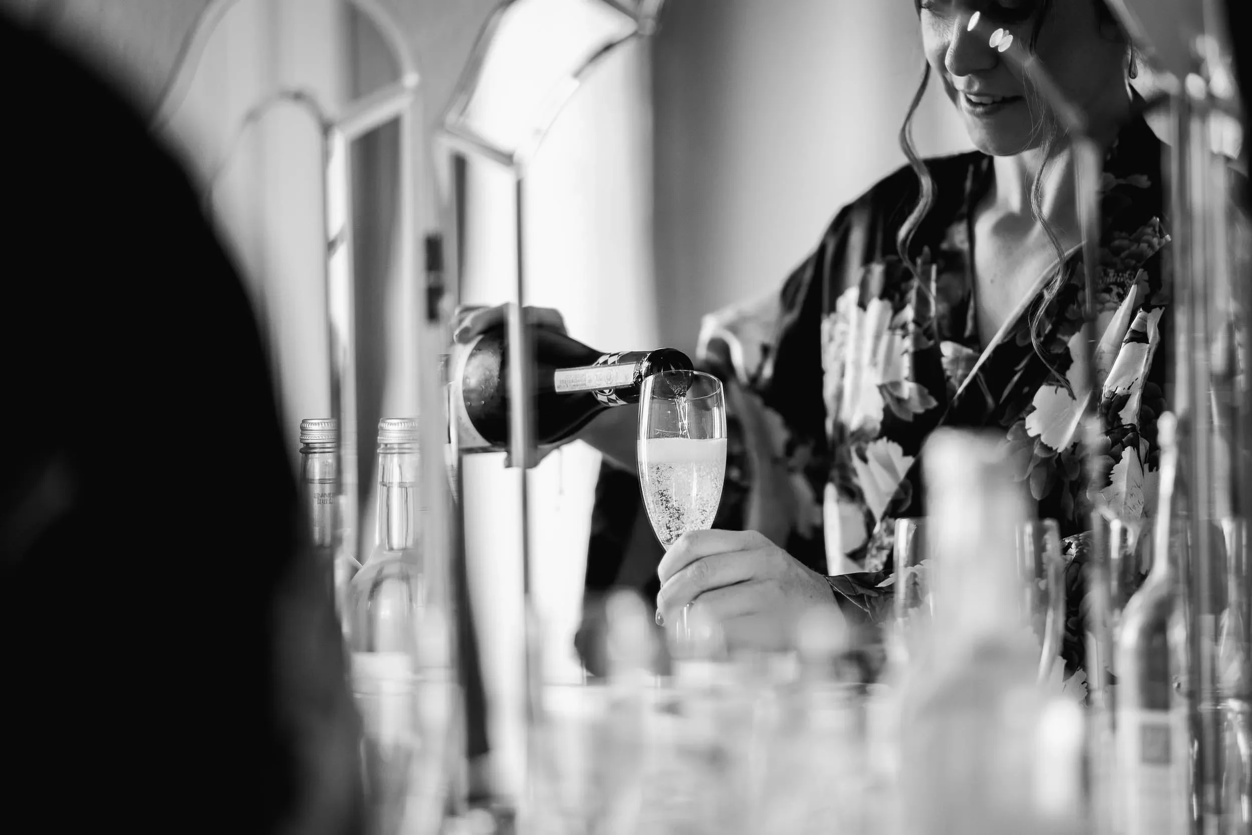 A woman pouring champagne into a flute at a bar, dressed in a floral robe, with bottles on the bar, in black and white.