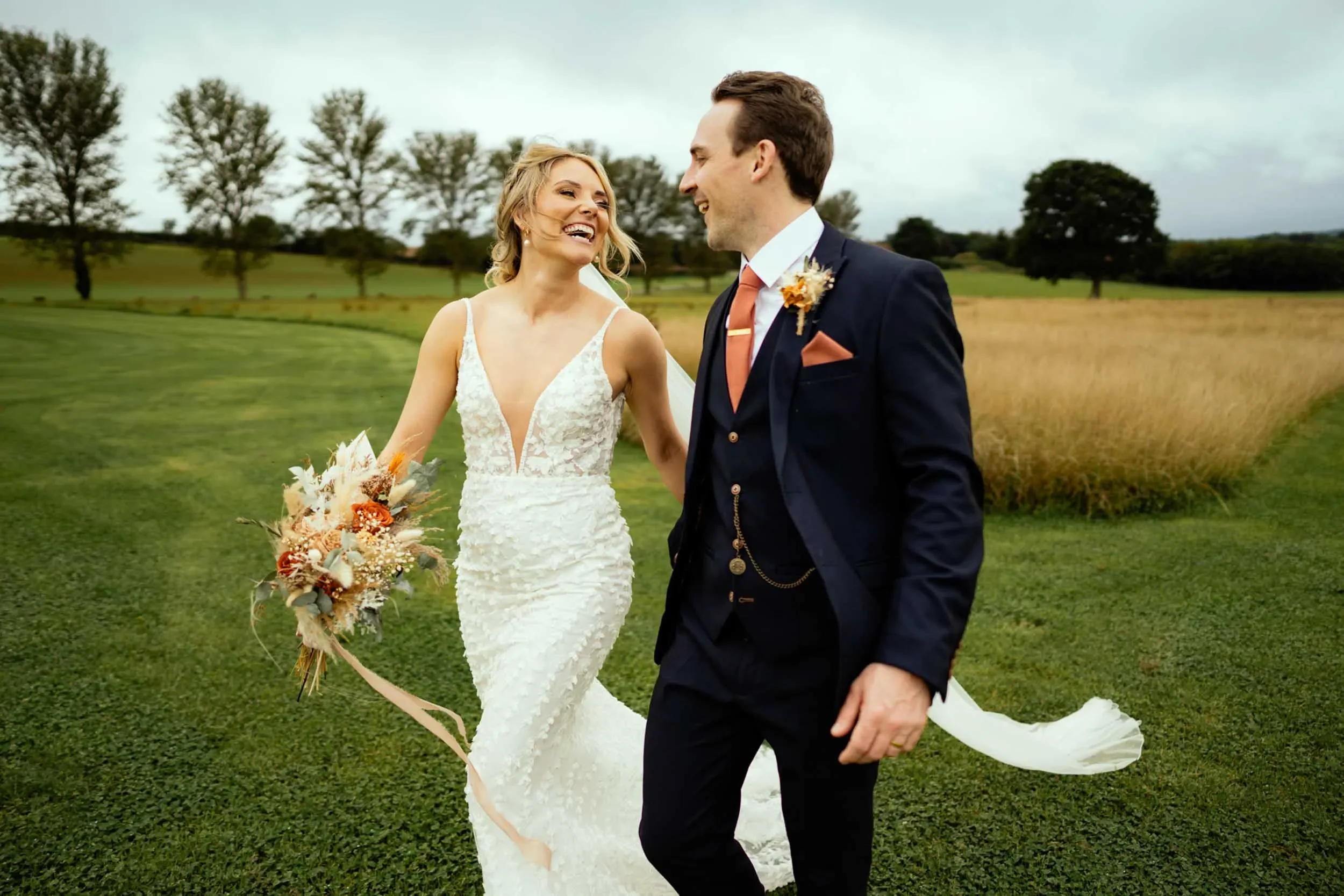 A happy bride and groom walking outdoors on a green field on a cloudy day, smiling and looking at each other.