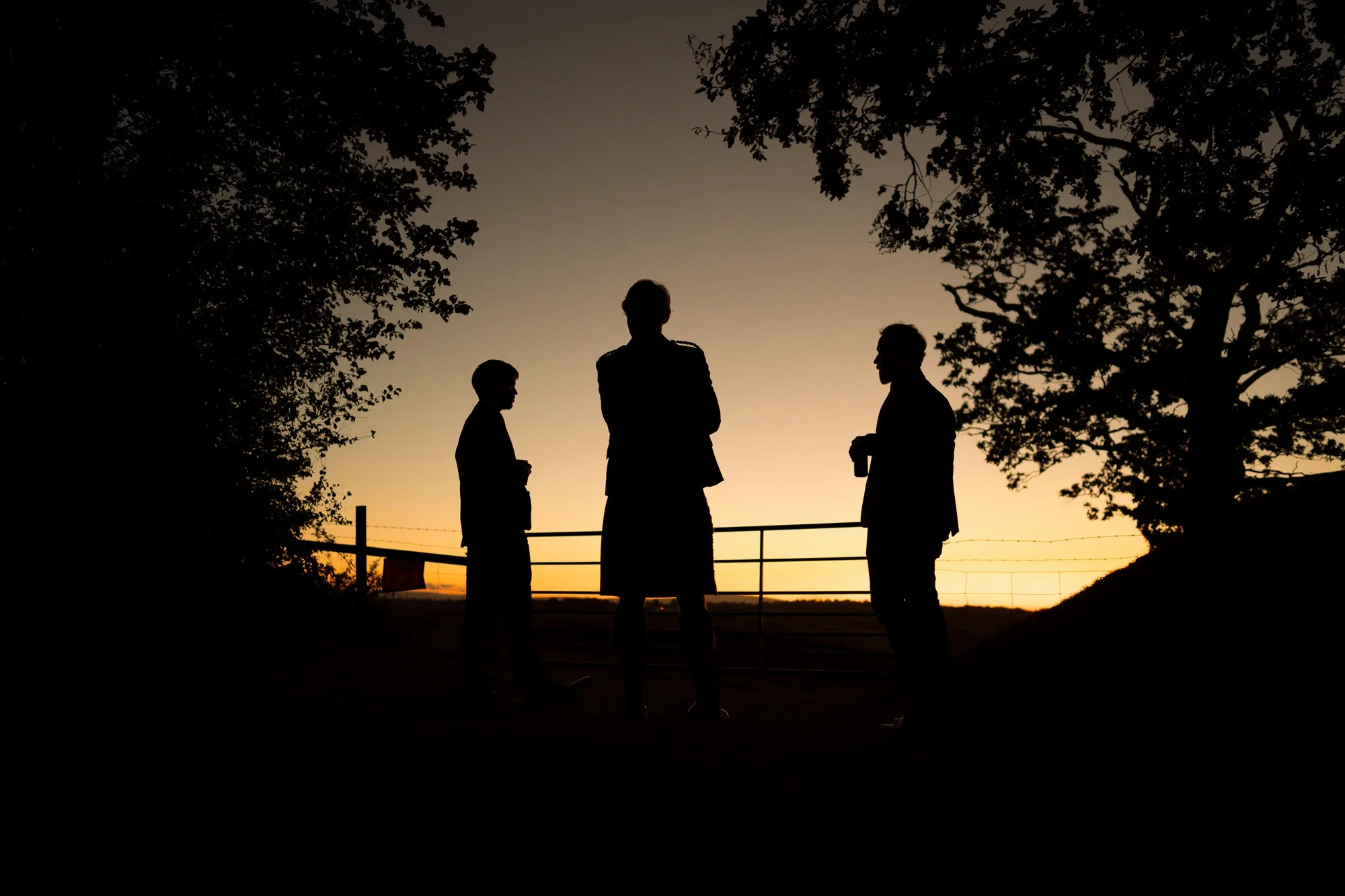 Silhouettes of three men and two women standing outdoors at sunset, with trees and a fence in the background.