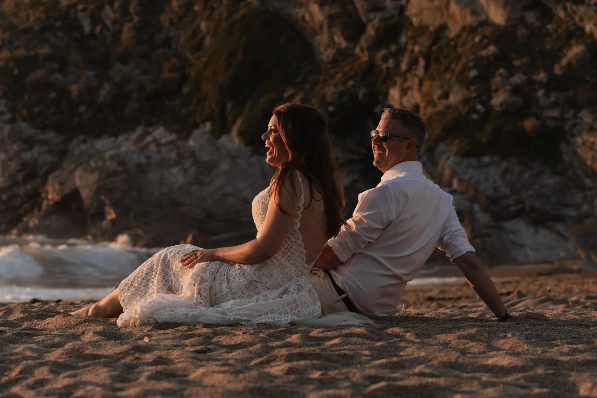 A couple sitting on a sandy beach during sunset, laughing and enjoying each other's company, with rocky cliffs in the background.