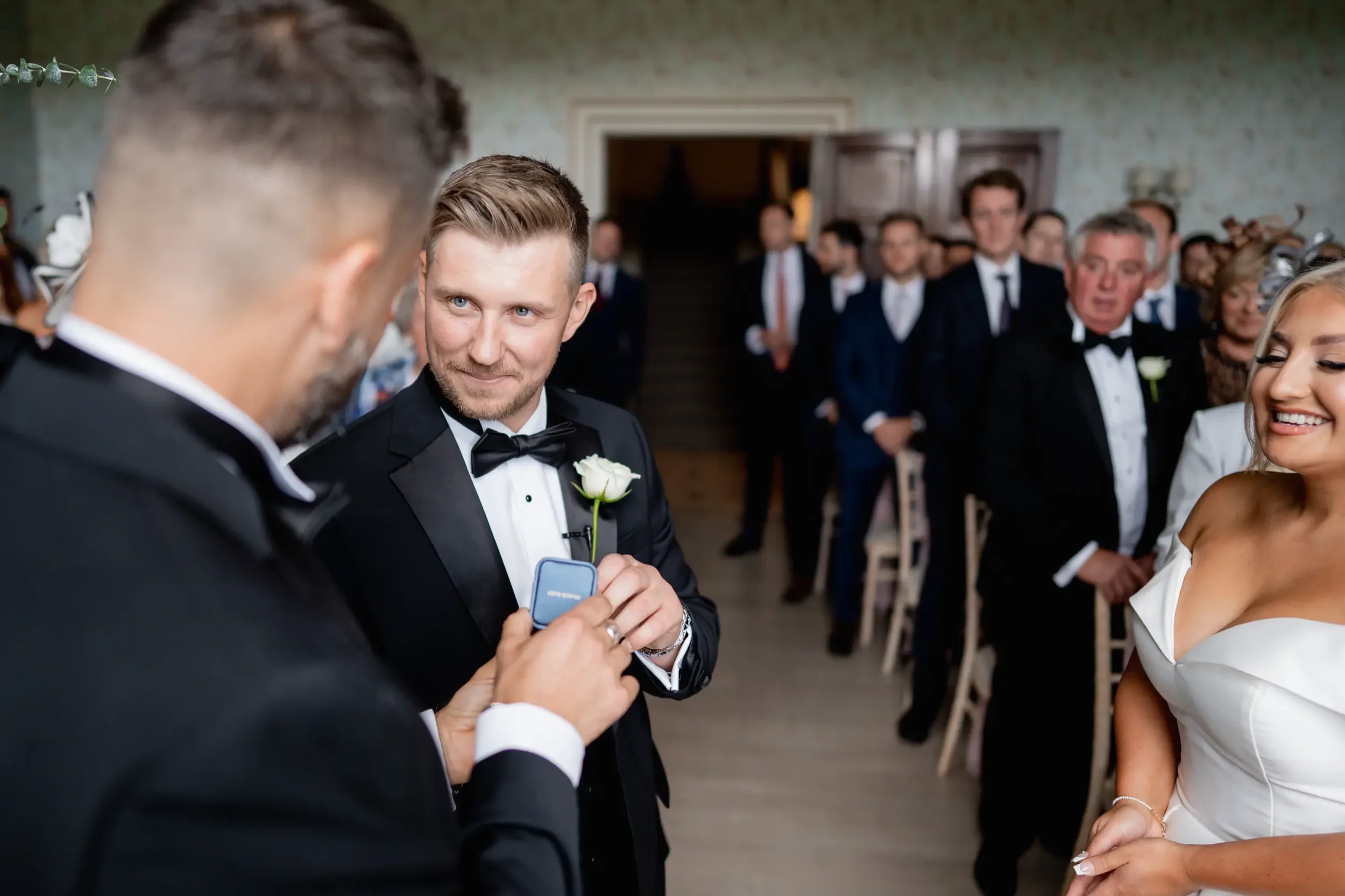 A man in a tuxedo with a white rose boutonniere is holding a ring box, while another man in a tuxedo looks on during a wedding ceremony with guests in the background.