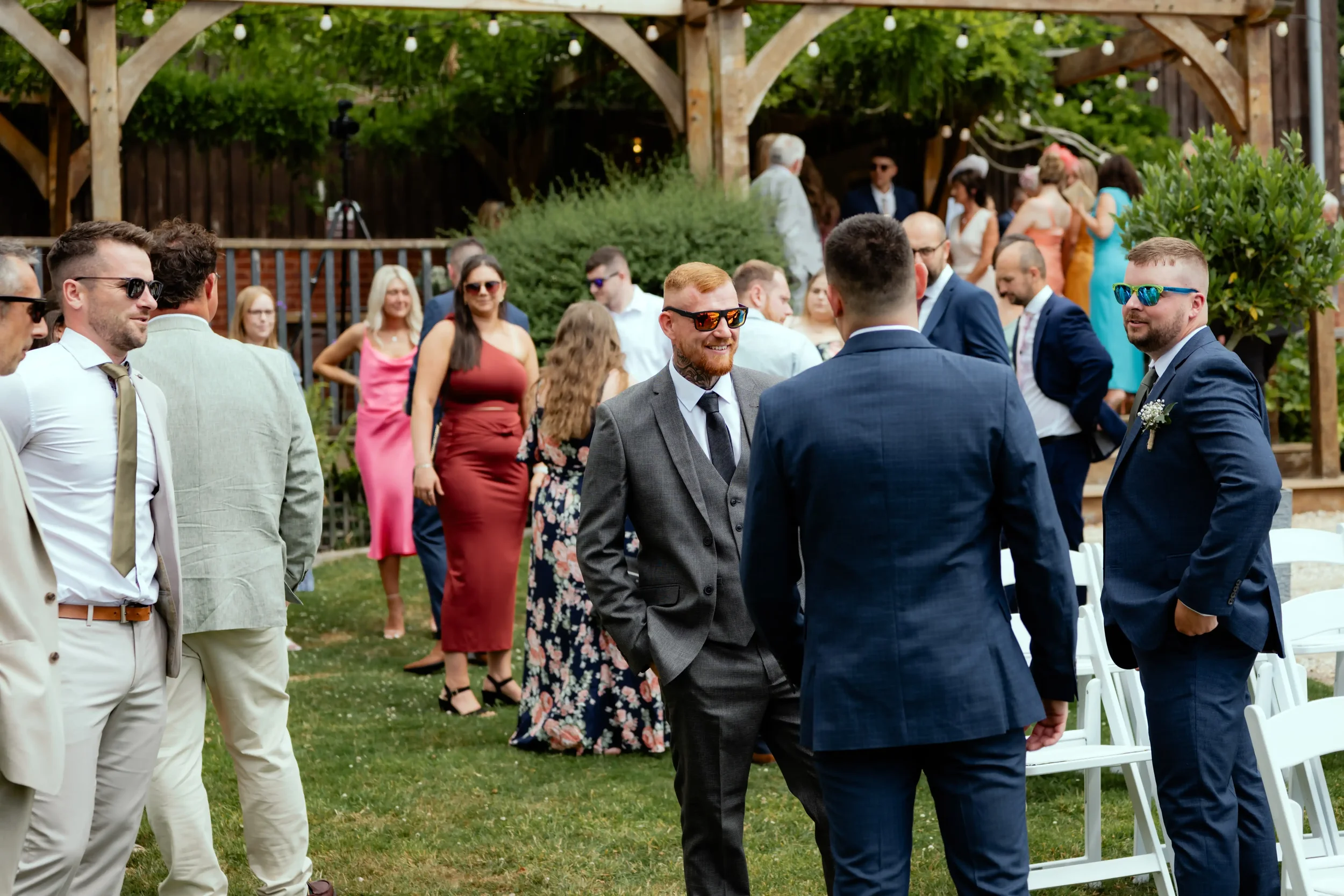 People dressed in formal attire socializing outdoors at a wedding reception or event, some wearing sunglasses, with a wooden pergola, string lights, and greenery in the background.