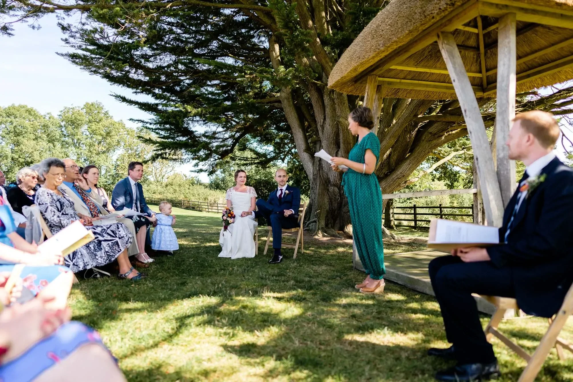 Outdoor wedding ceremony under a large tree. The bride and groom are seated, listening to a woman reading from a paper. Guests are seated on both sides, watching the scene. The setting is bright and sunny.