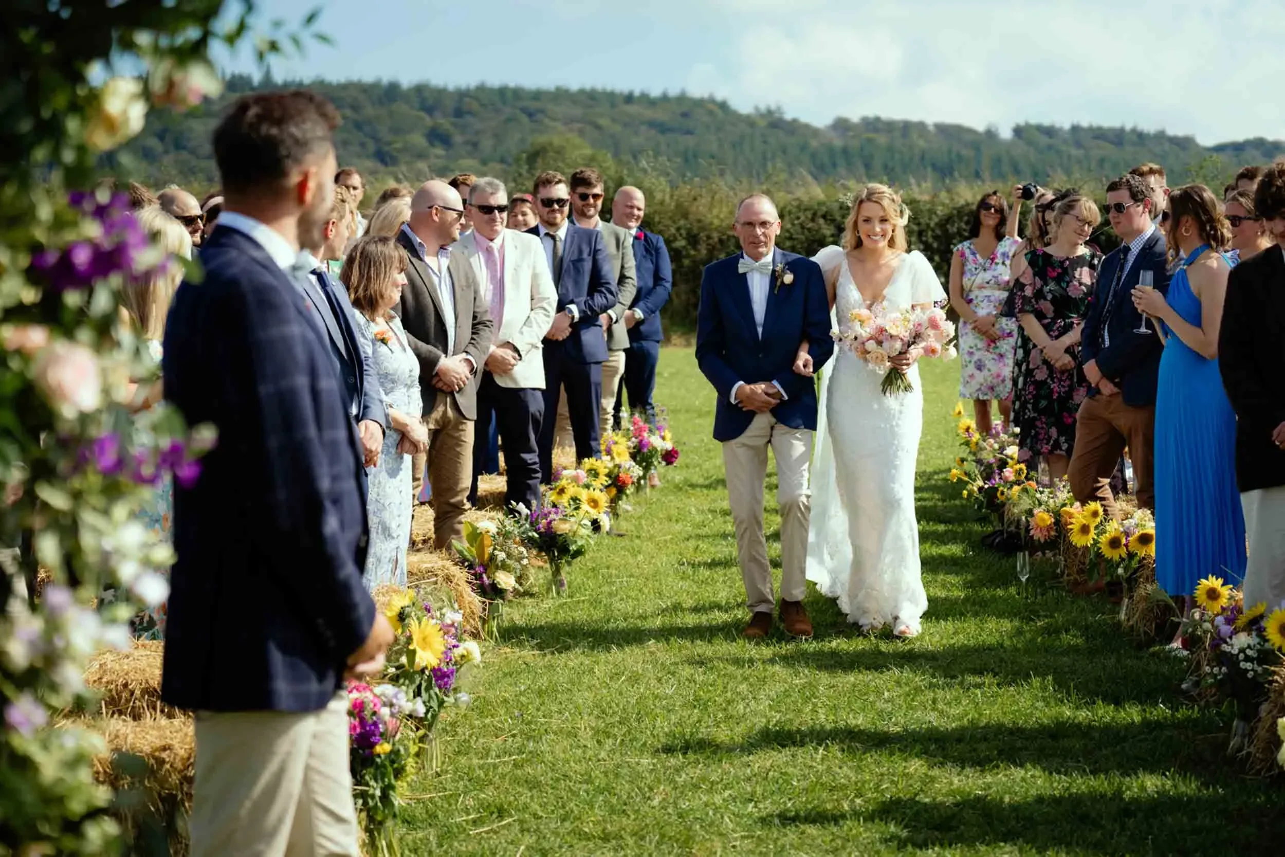 Bride walking down aisle with her father during outdoor wedding ceremony, surrounded by friends and family on a sunny day.