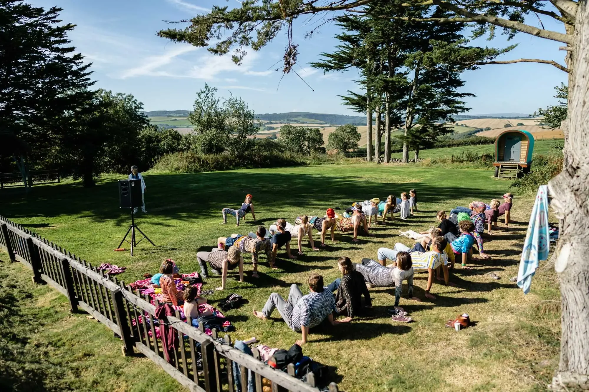 People participating in outdoor yoga class in a park on a sunny day with trees and hills in the background.