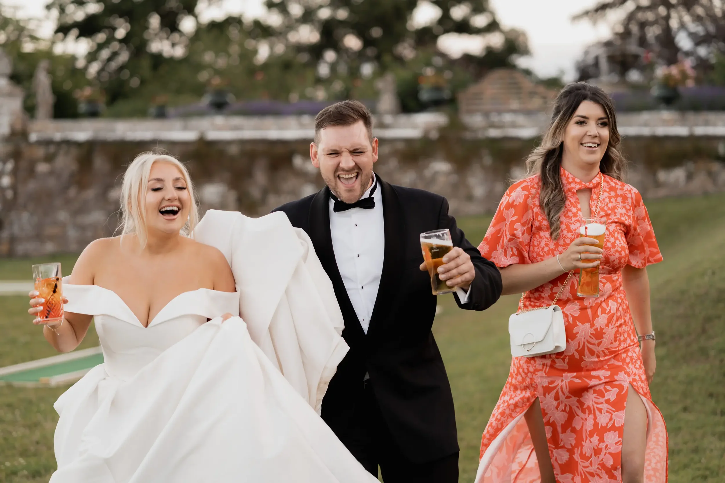 A wedding celebration outdoors with a bride in a white gown, a groom in a tuxedo, and a woman in a floral dress holding drinks, all smiling and enjoying the moment.
