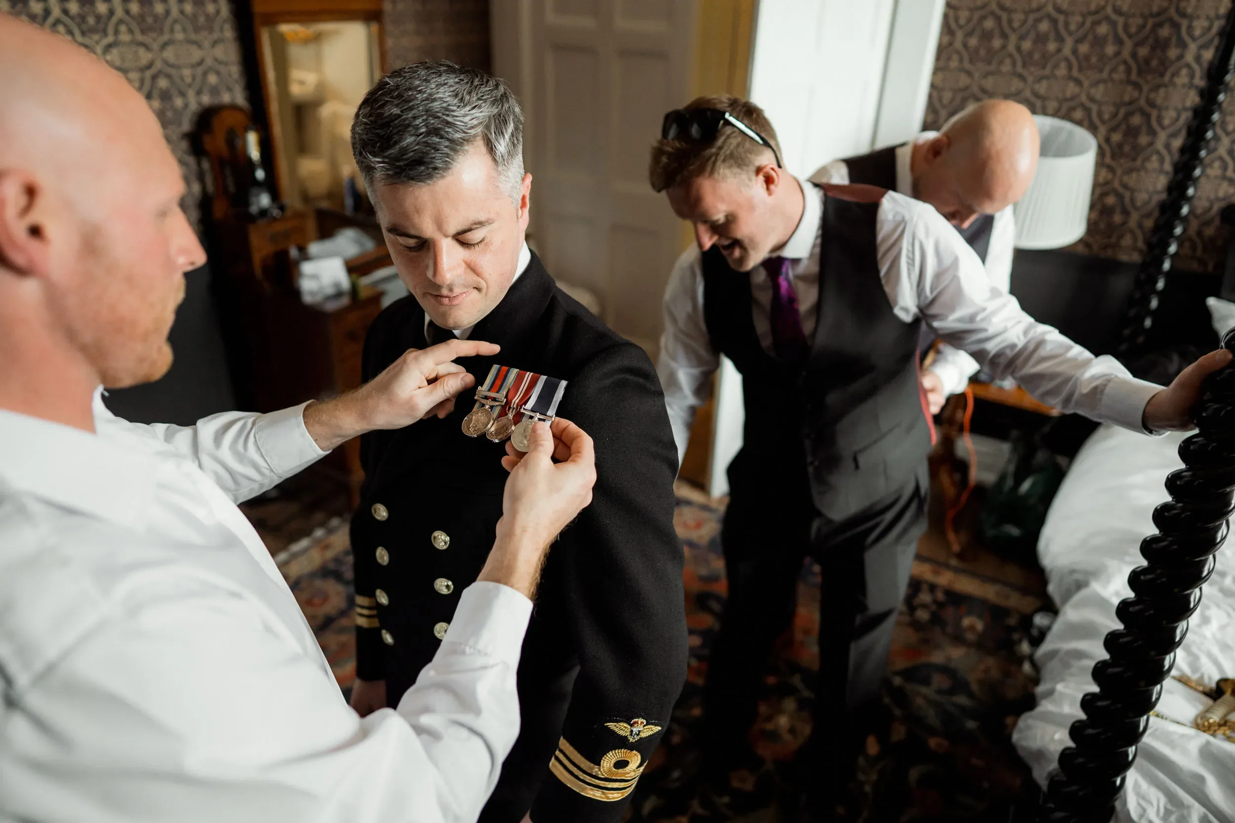 Four men in formal attire in a hotel room, one man in a military uniform receiving medals from another man, while another man looks on.