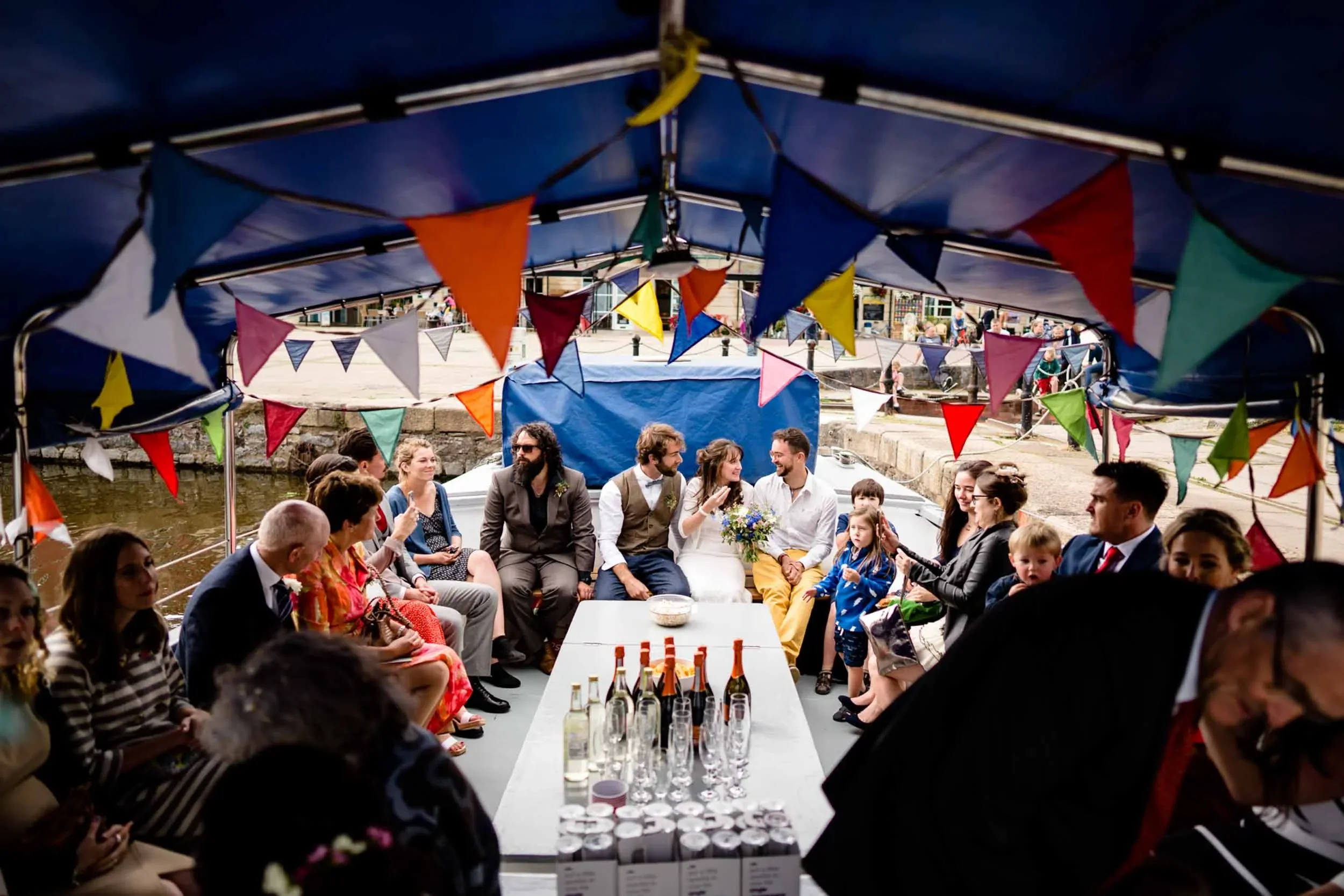 A boat decorated with colorful bunting is filled with guests celebrating a wedding, with the bride and groom seated in the middle and tables with drinks inside the boat.