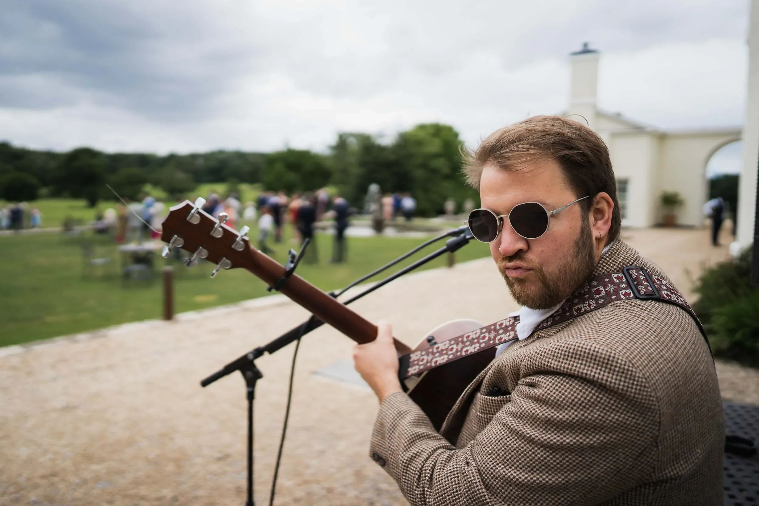 A man wearing sunglasses and a brown checkered jacket playing an acoustic guitar outdoors at a park or garden. There are people in the background and a white building with an arched entrance.