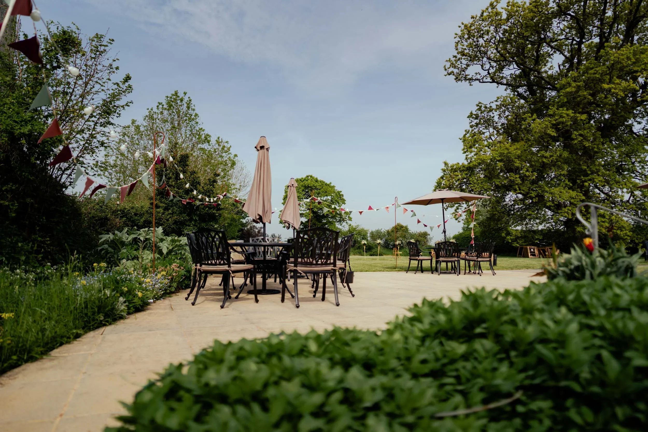 An outdoor patio with round tables and chairs, decorated with umbrellas and string banners, surrounded by trees and greenery.