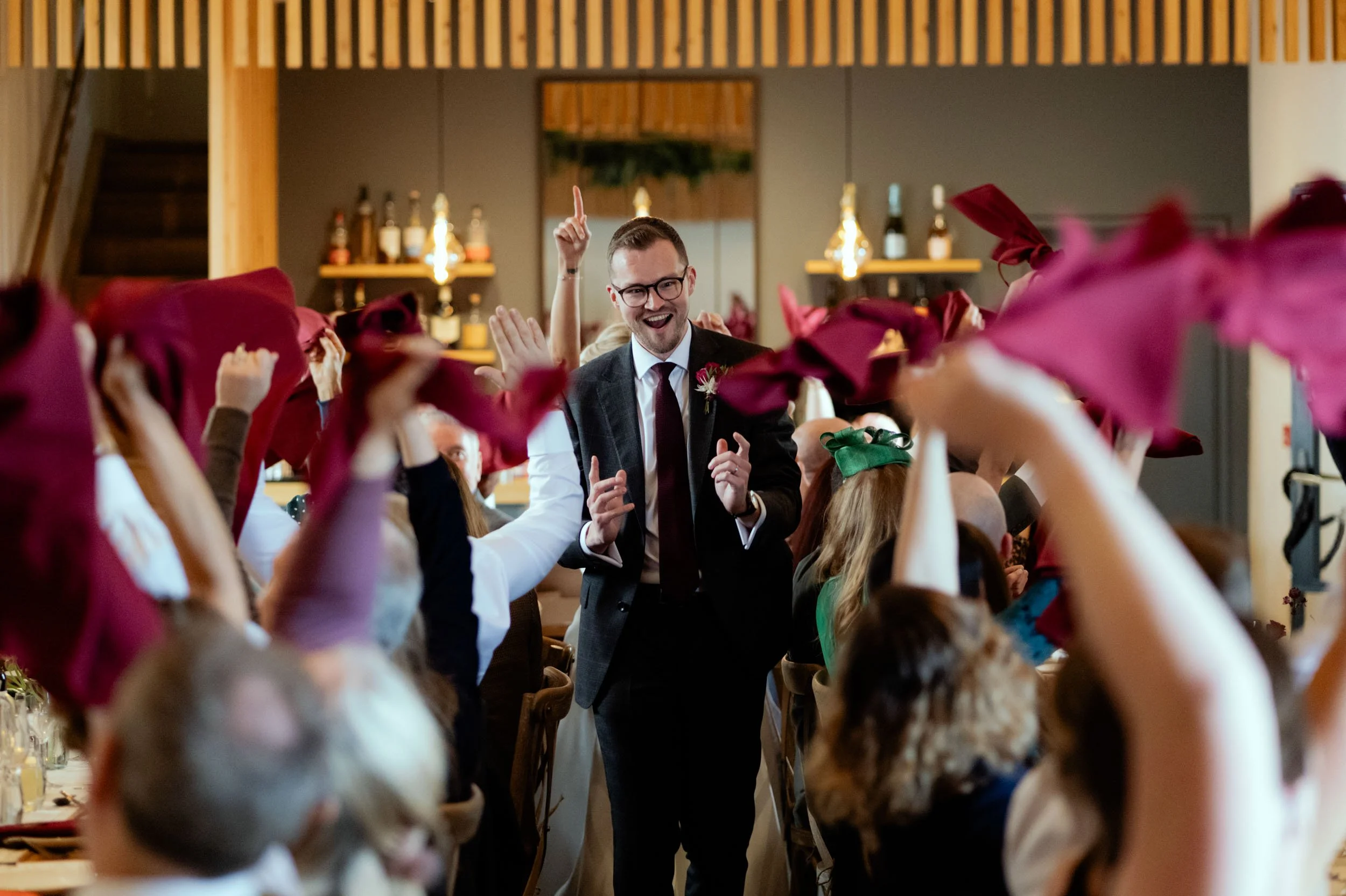 A man in a suit and glasses dancing with a joyful expression at a celebration, surrounded by guests with arms raised holding red napkins or cloths, in a decorated indoor venue.