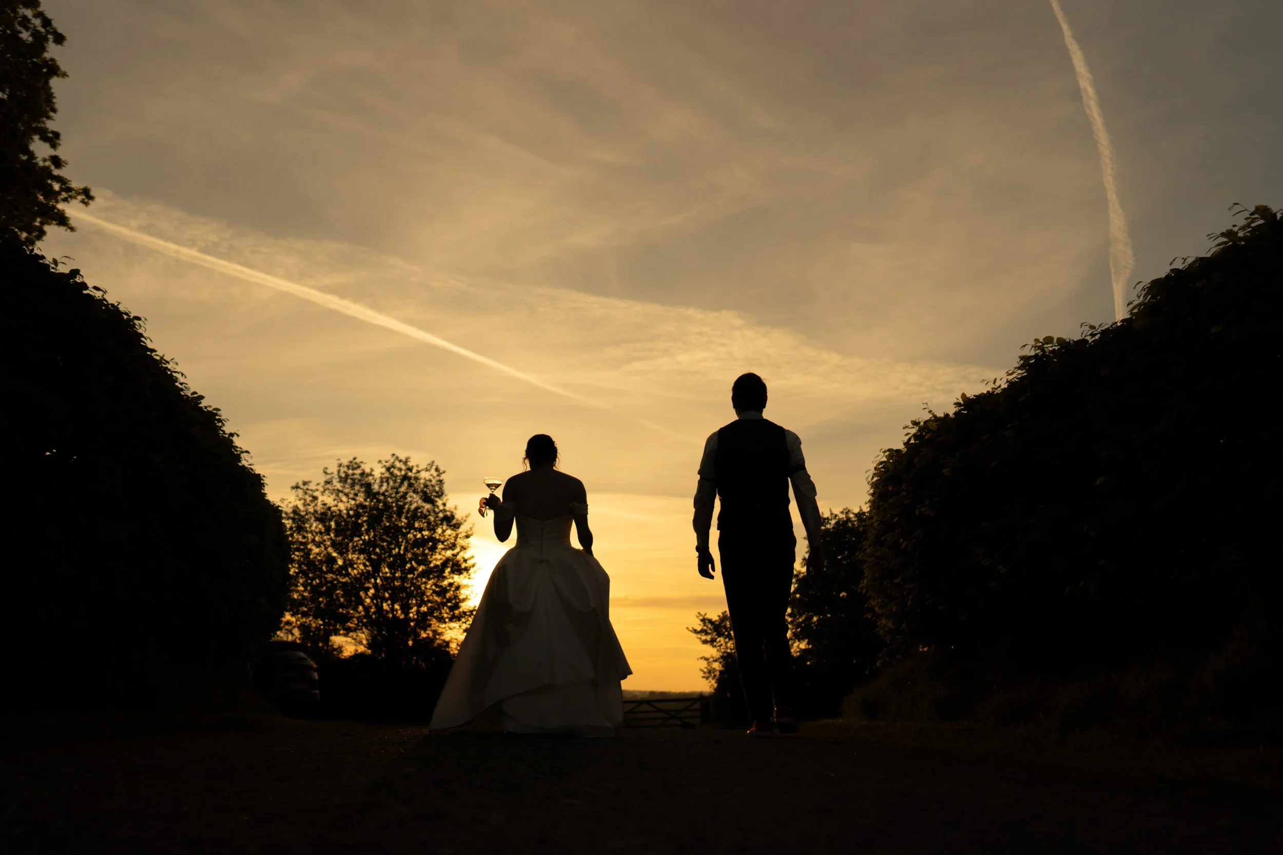 Silhouettes of a bride holding a glass of wine and a groom walking during sunset.
