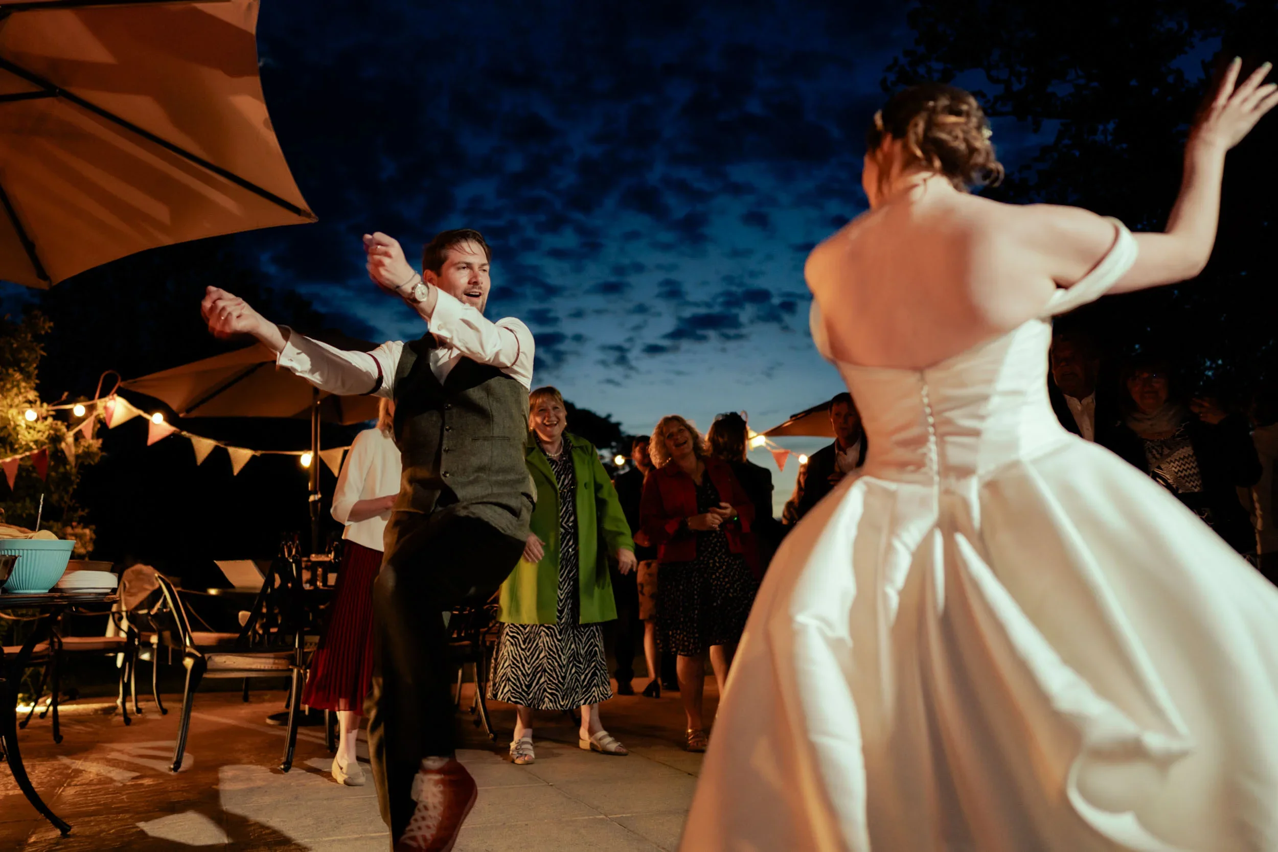 Couple dancing at an outdoor wedding reception at night, with guests watching and smiling in the background, under string lights and umbrellas.