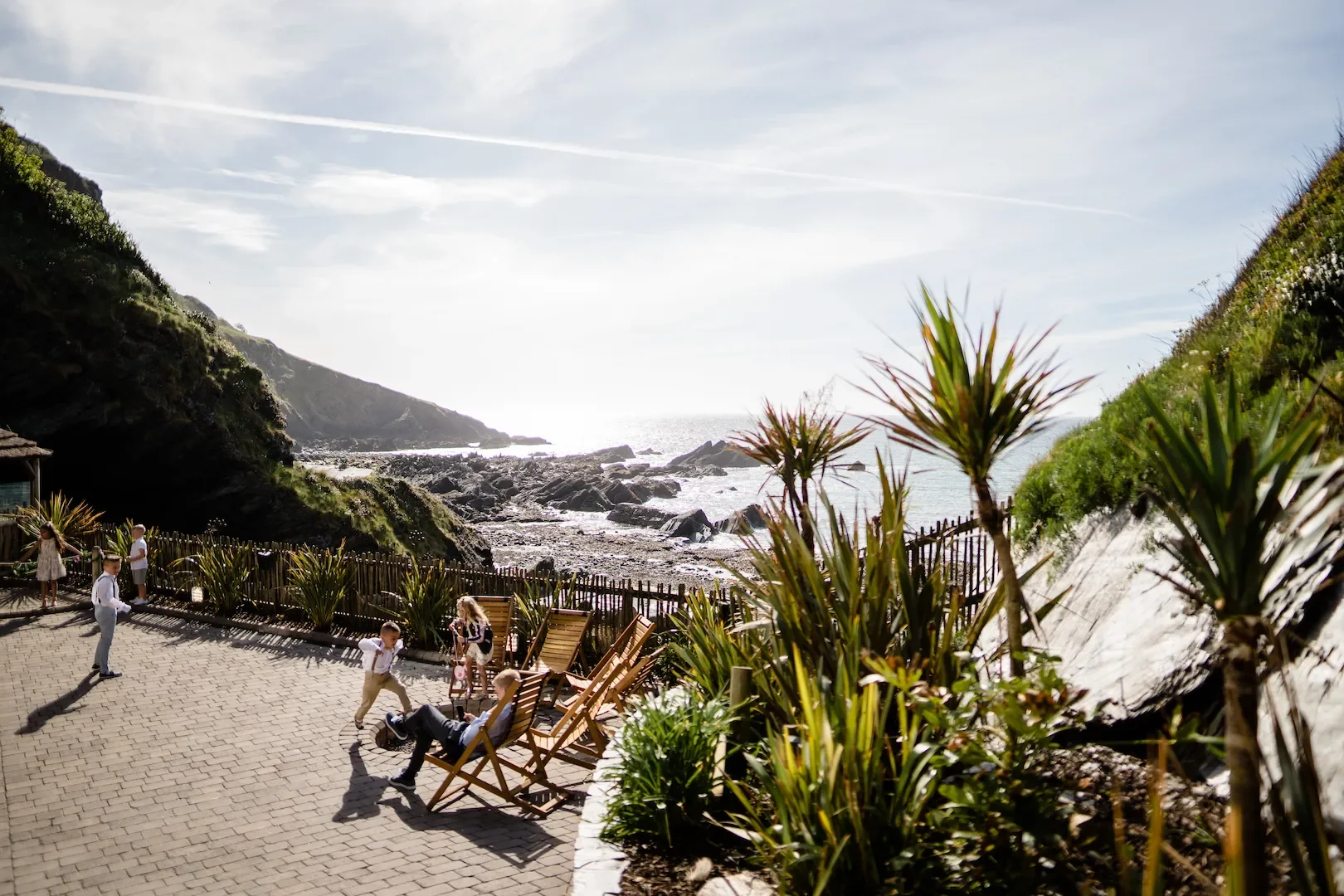 People relaxing and children playing on a paved outdoor area with wooden lounge chairs, surrounded by green plants, overlooking rocky coastline and ocean in the background under a partly cloudy sky.
