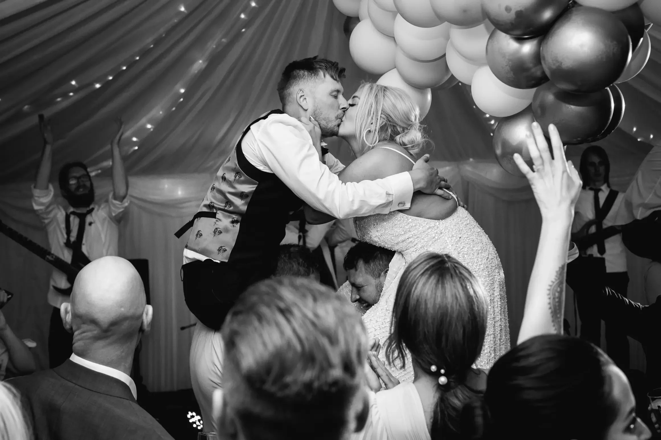A black-and-white photo of a wedding celebration with a couple kissing, supported by the groom in a playful lift, surrounded by guests and musicians under a decorated tent.