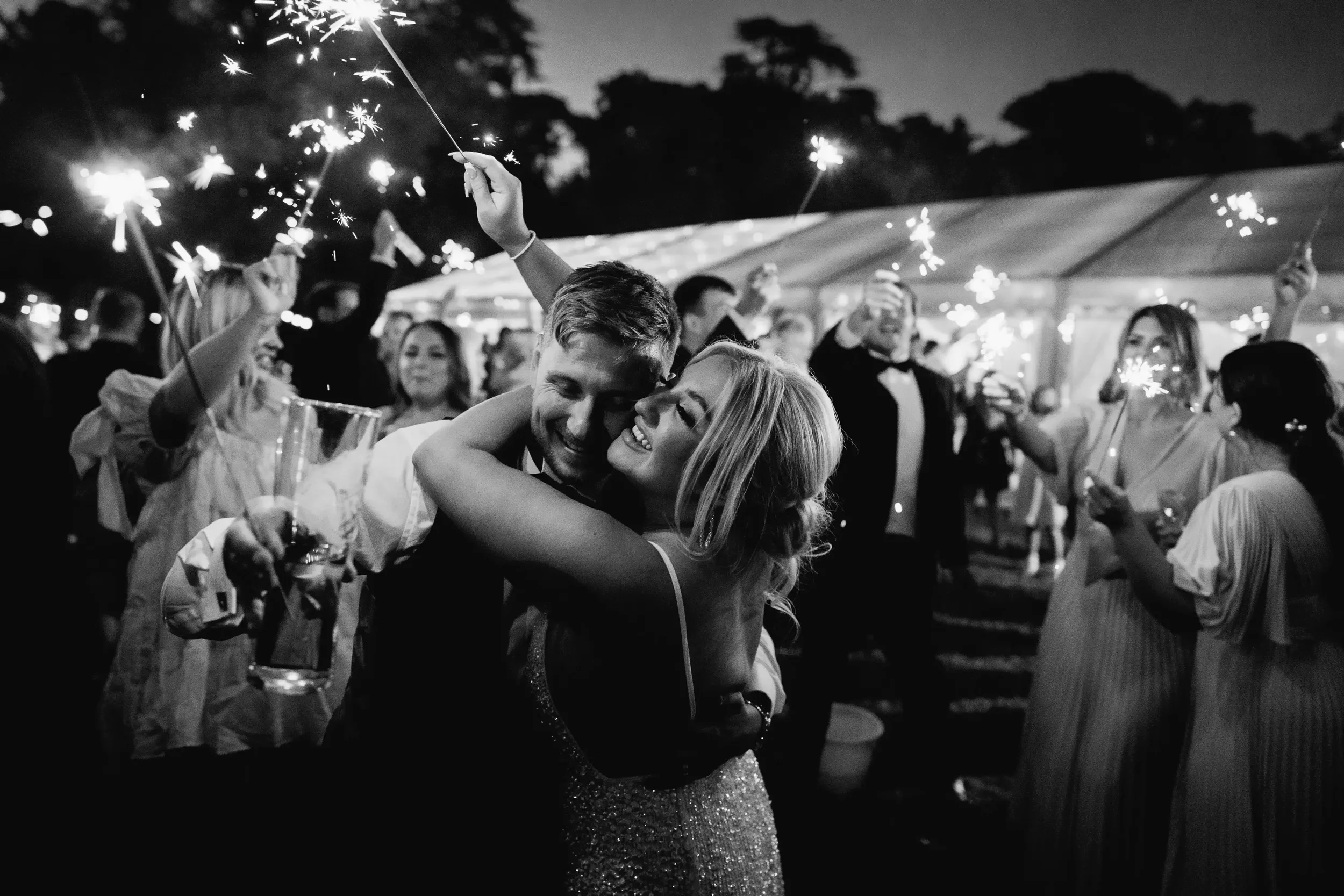 A black and white photo of a wedding celebration outdoors at night, with a group of people holding sparklers. In the center, a happy couple is dancing and embracing, smiling joyfully.