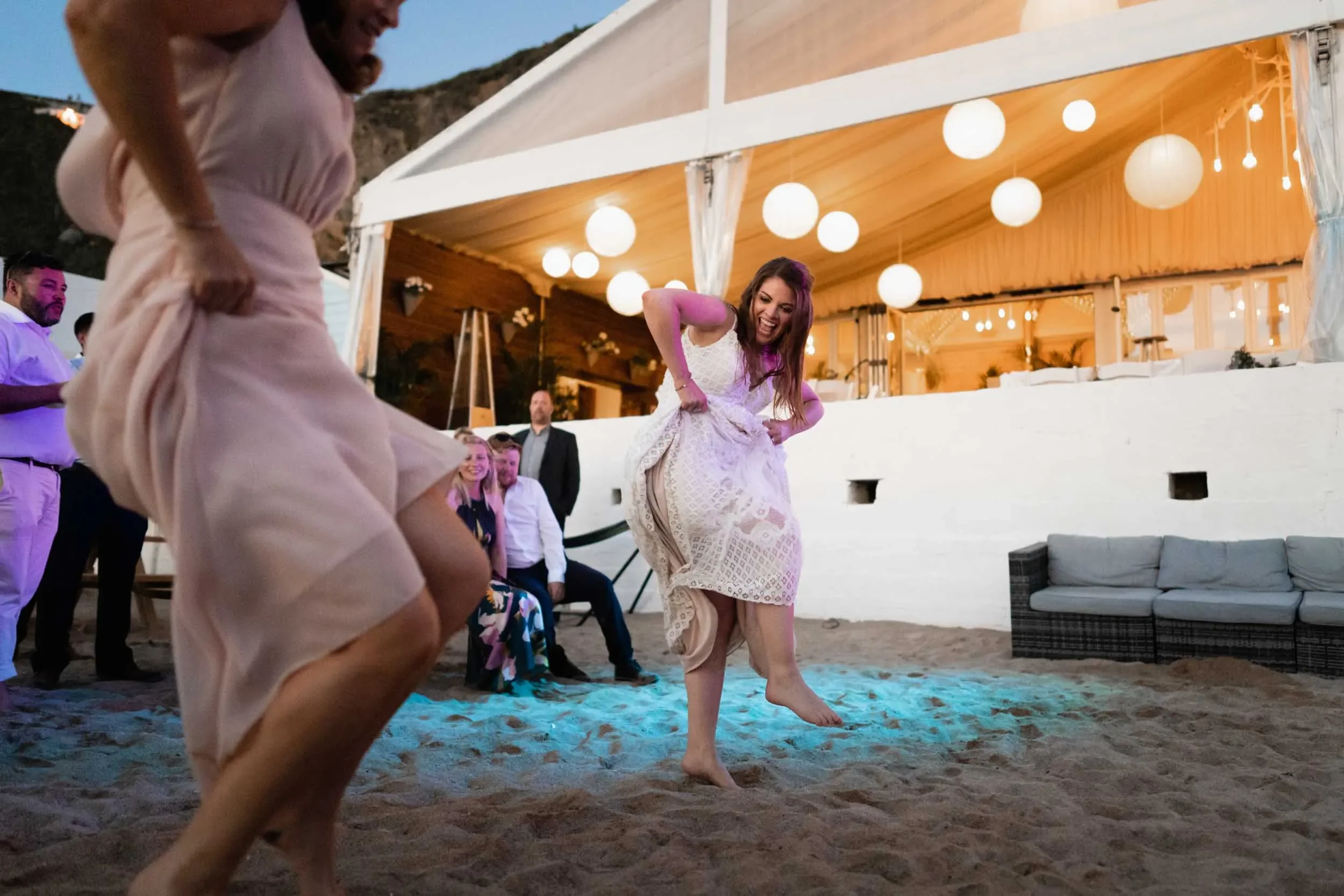 Two women dancing on a sandy outdoor area during a celebration, with one woman in a white dress raising her dress, and several people watching in the background under a decorated outdoor pavilion with hanging white lanterns.