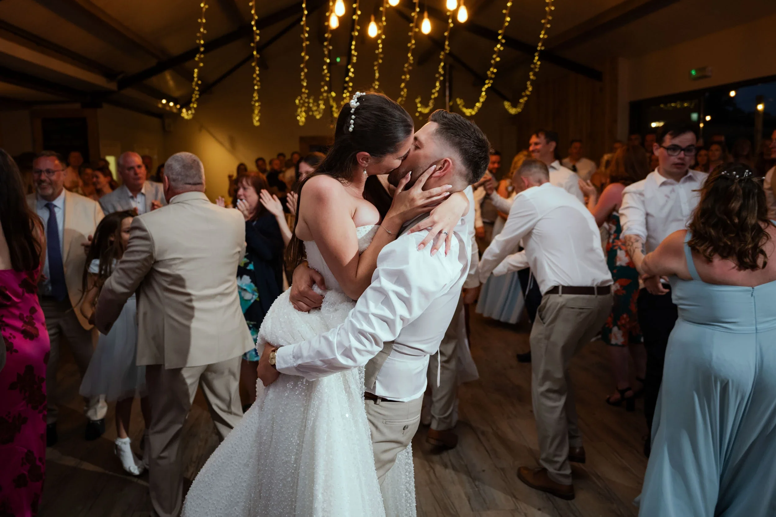 A bride and groom kiss while dancing at their wedding reception, surrounded by guests on a dance floor decorated with hanging lights.