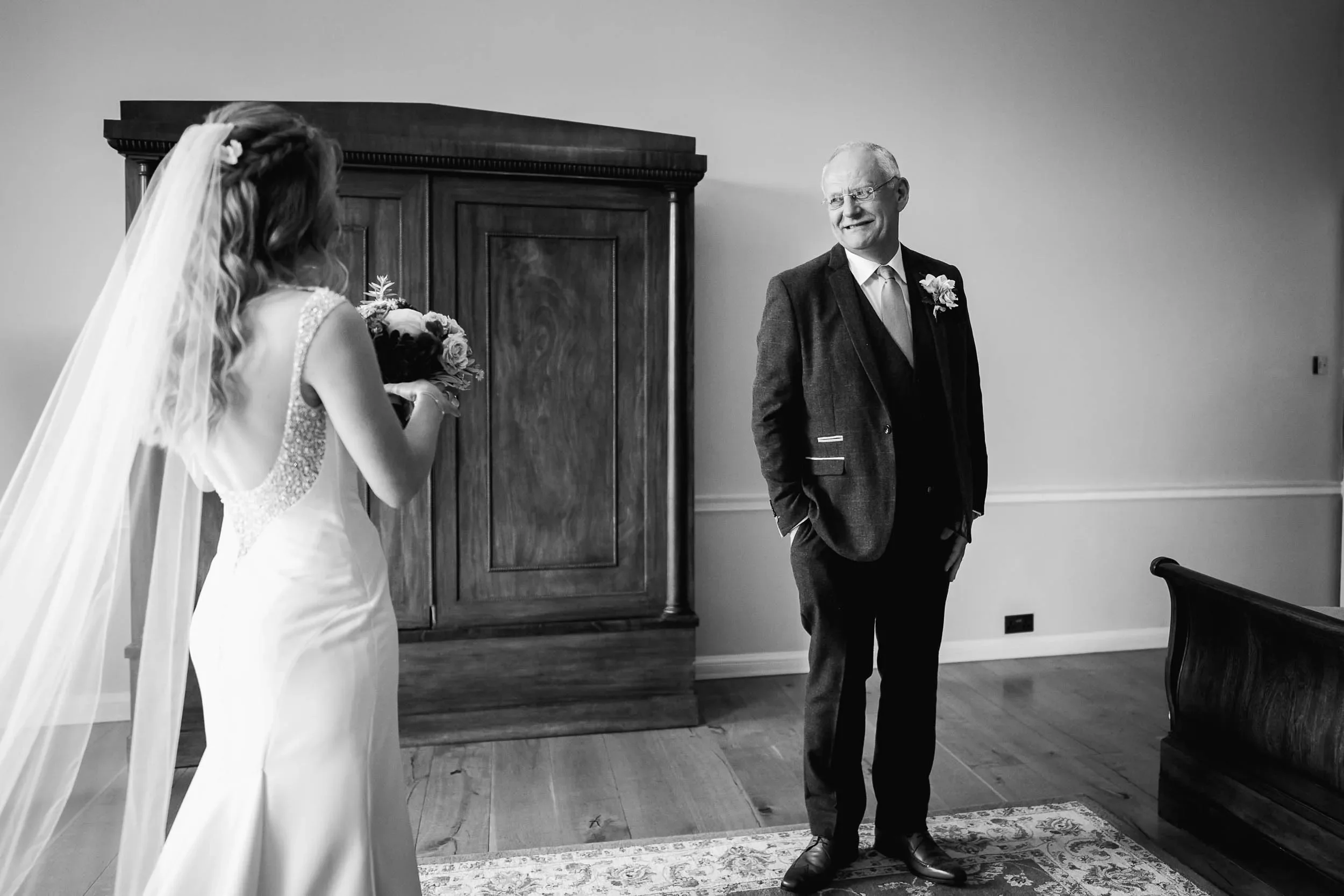Black-and-white photo of a bride holding flowers and a groom standing in a room, looking at her, with a wooden cabinet and window in the background.
