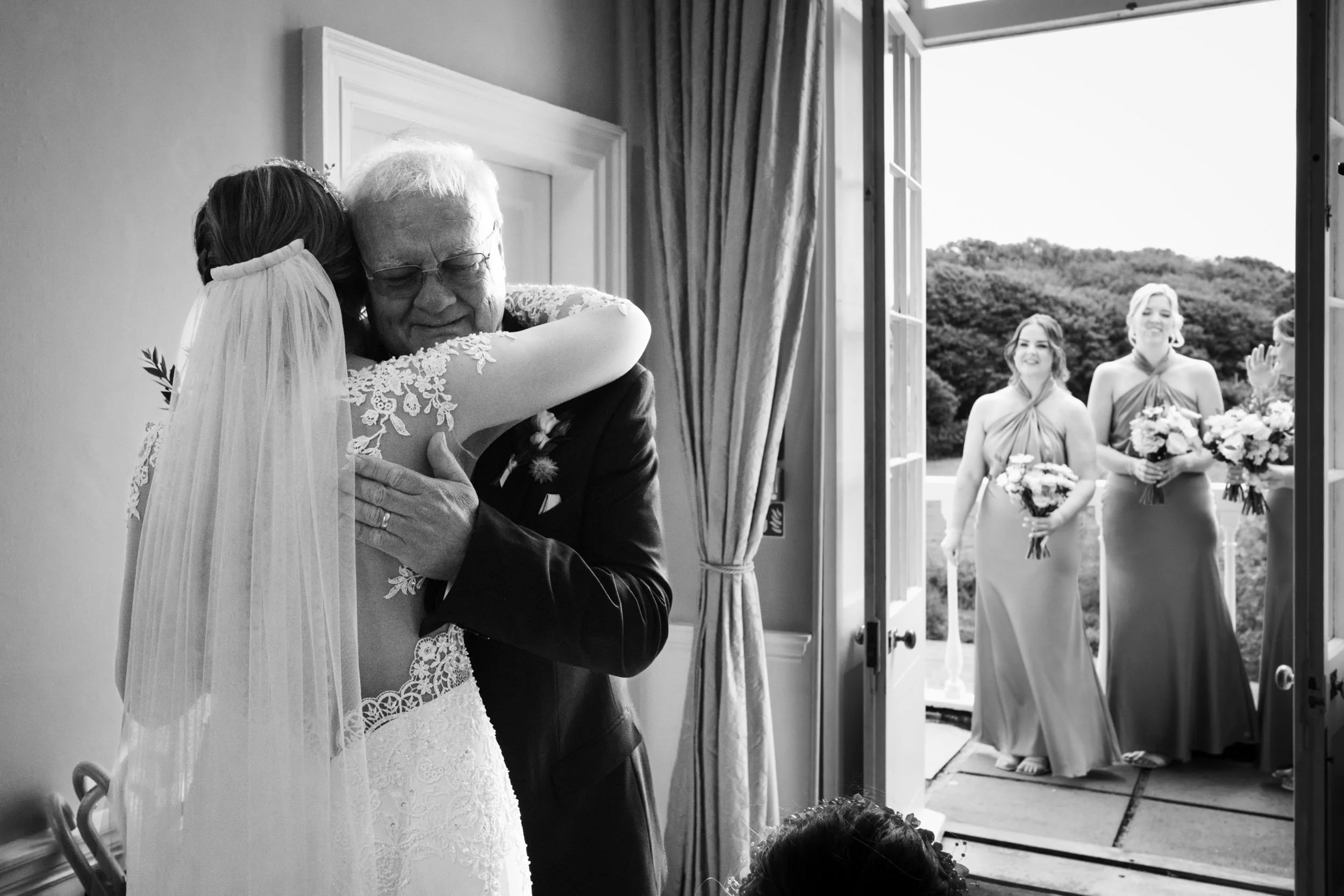 A bride hugging an elderly man, likely her father, in a wedding dress inside a room. Outside, three bridesmaids are standing at the open door holding bouquets, smiling and looking toward the inside.