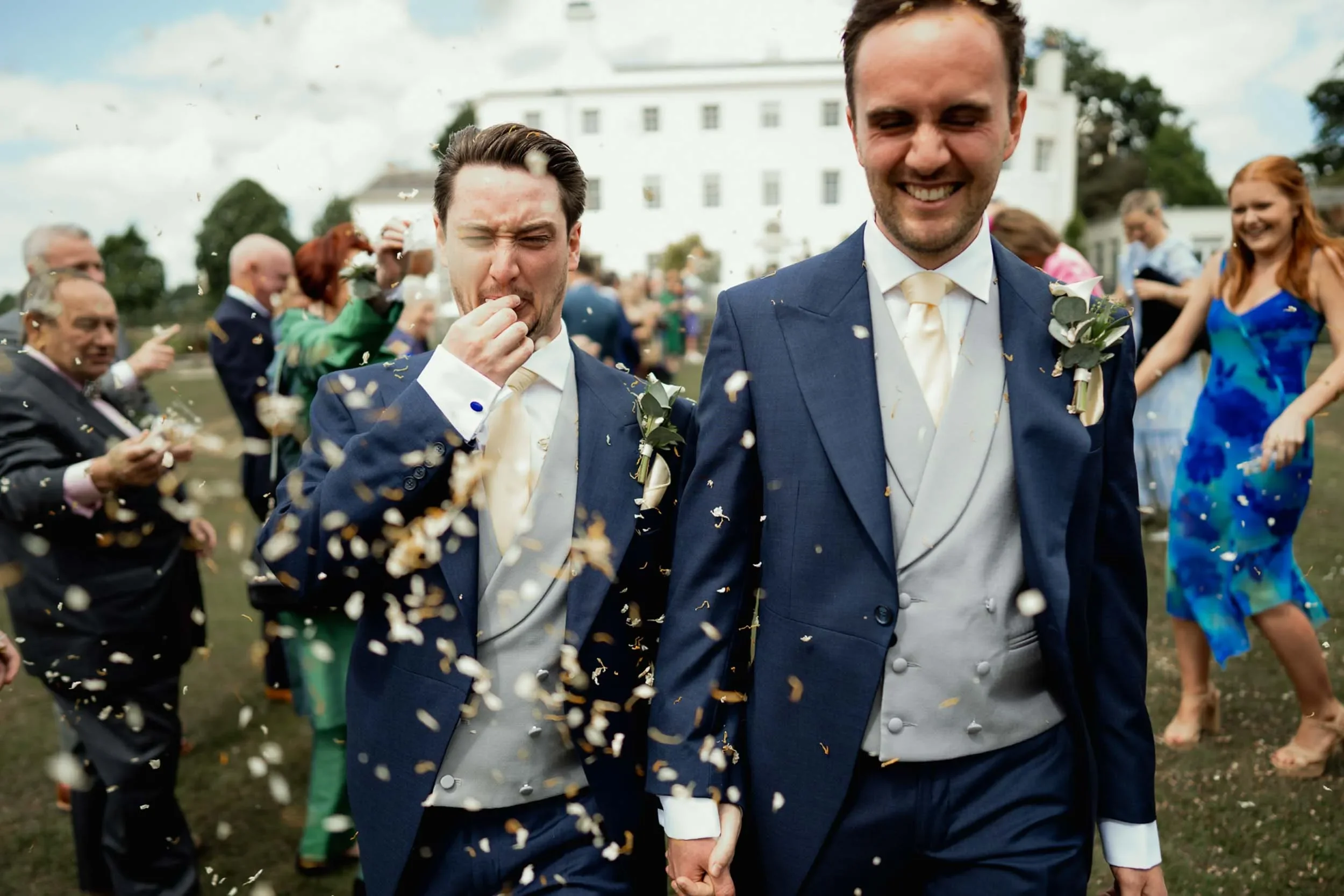 Two men wearing blue suits and vests, holding hands, smiling and celebrating with guests in the background outside a large white building, during a wedding celebration with confetti in the air.