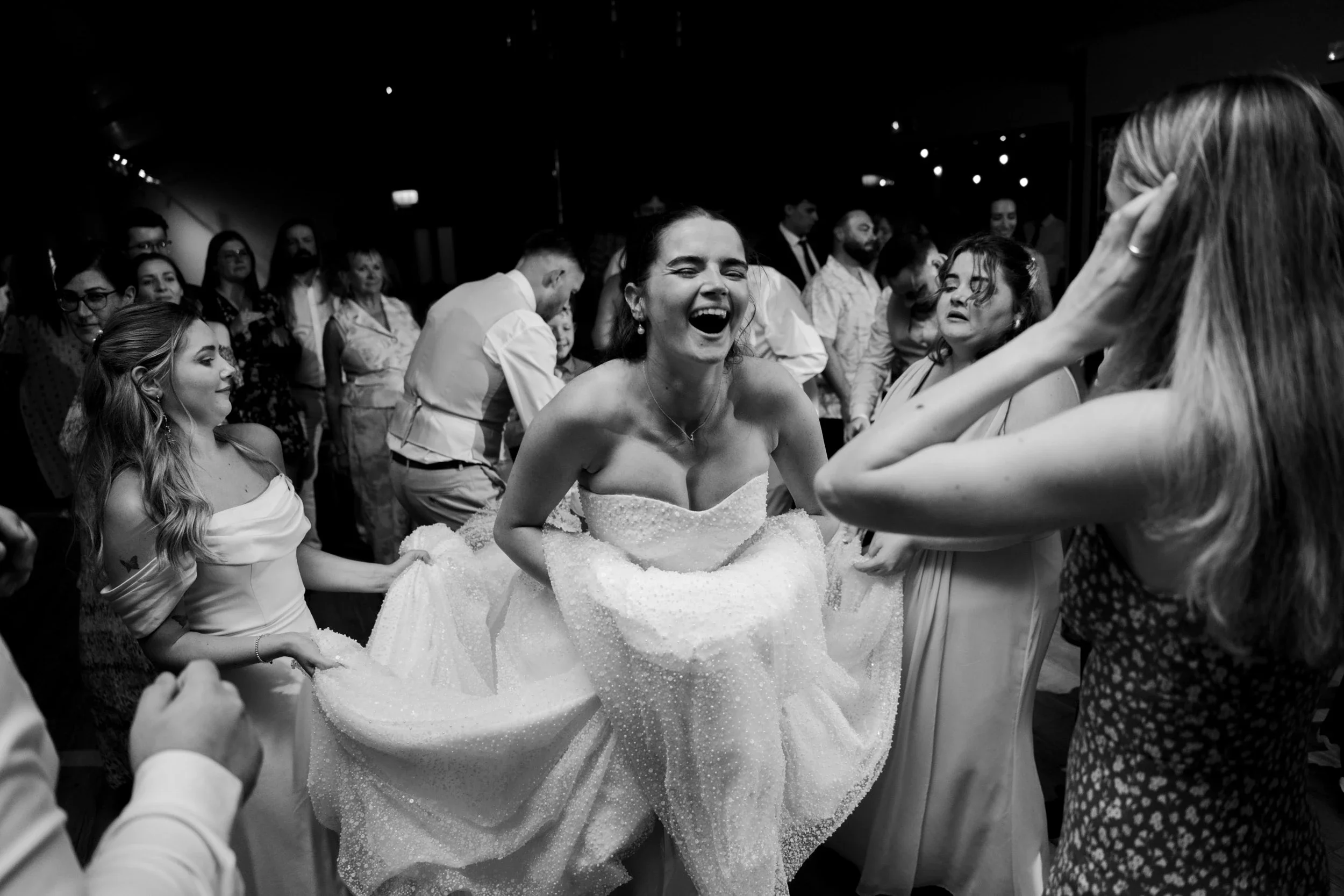 A bride laughing joyfully at her wedding reception as she is lifted in a chair by friends or family, surrounded by guests.