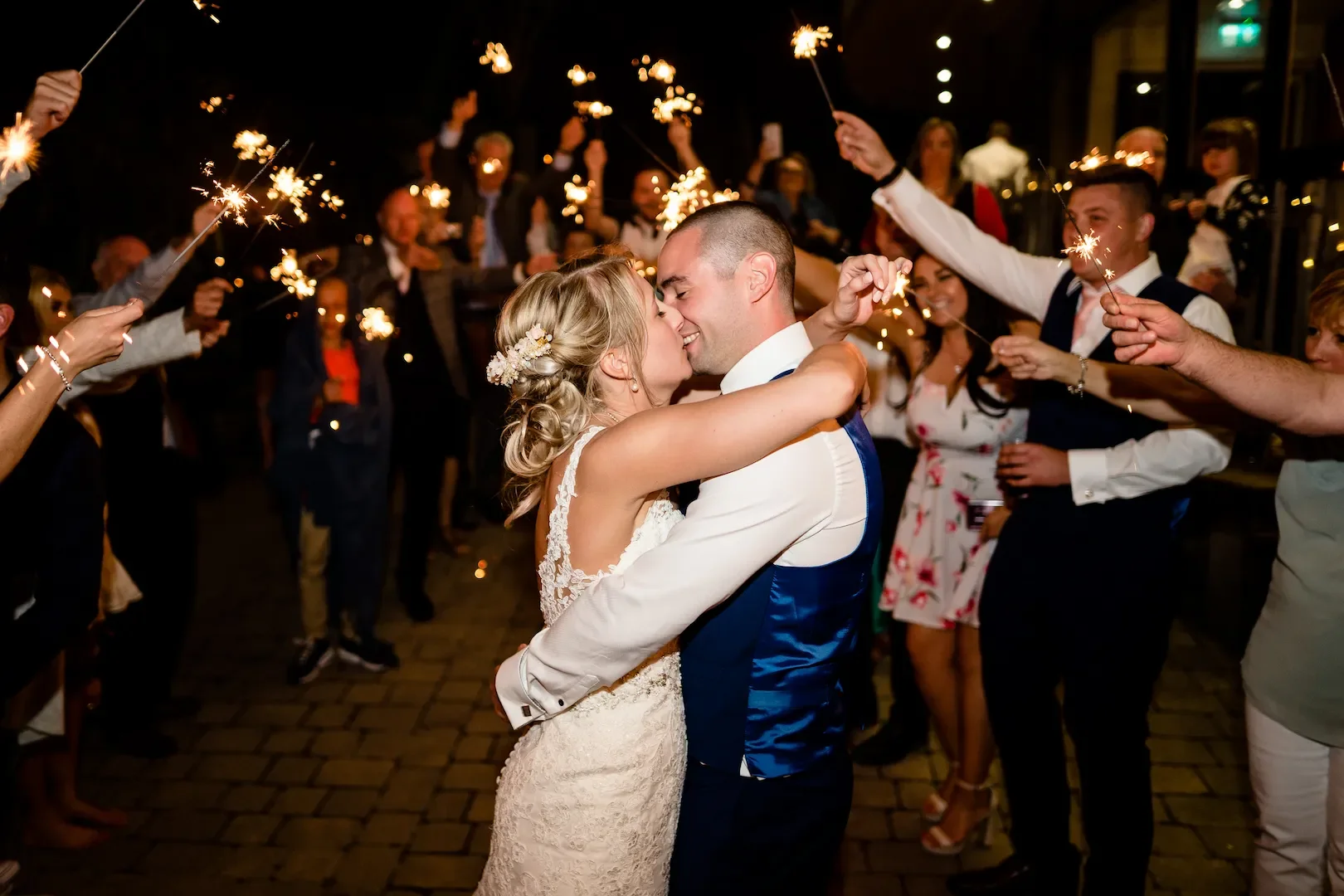 A newlywed couple embraces during their wedding celebration at night, surrounded by friends and family holding sparklers.