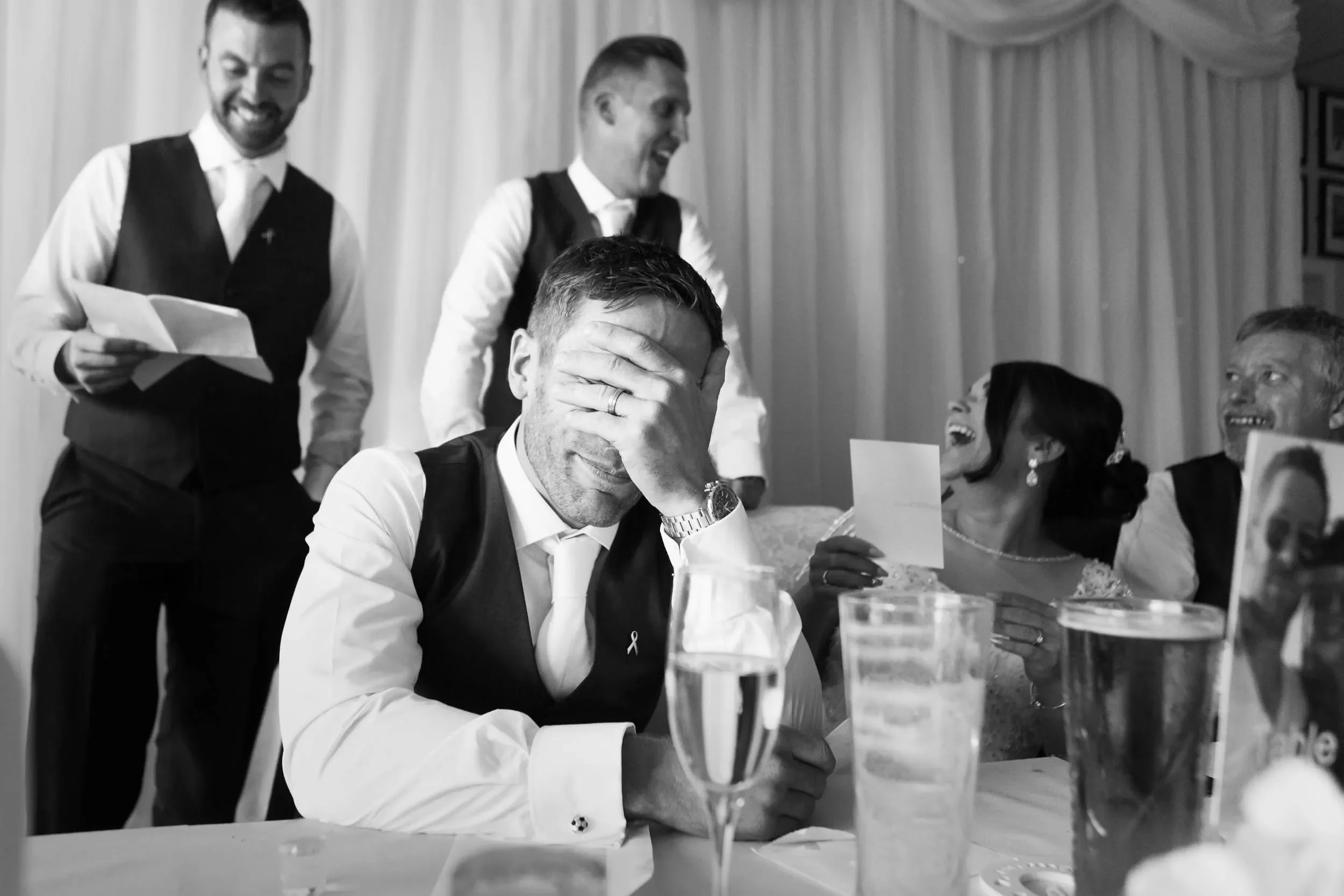 A group of people at a wedding celebration. The man in the center has his hand covering his face, appearing emotional or overwhelmed. The other individuals around him are smiling and laughing, with some holding drinks and greeting cards, indicating a