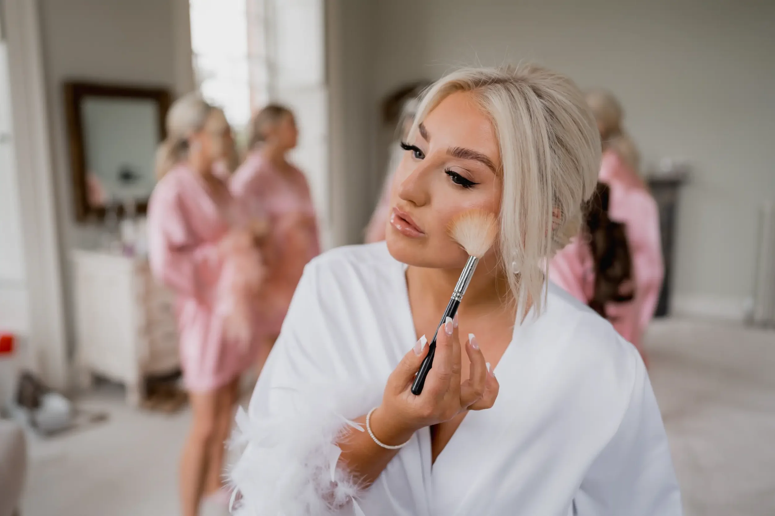 Bride applying makeup with a brush in a room with her bridesmaids in pink robes in the background.